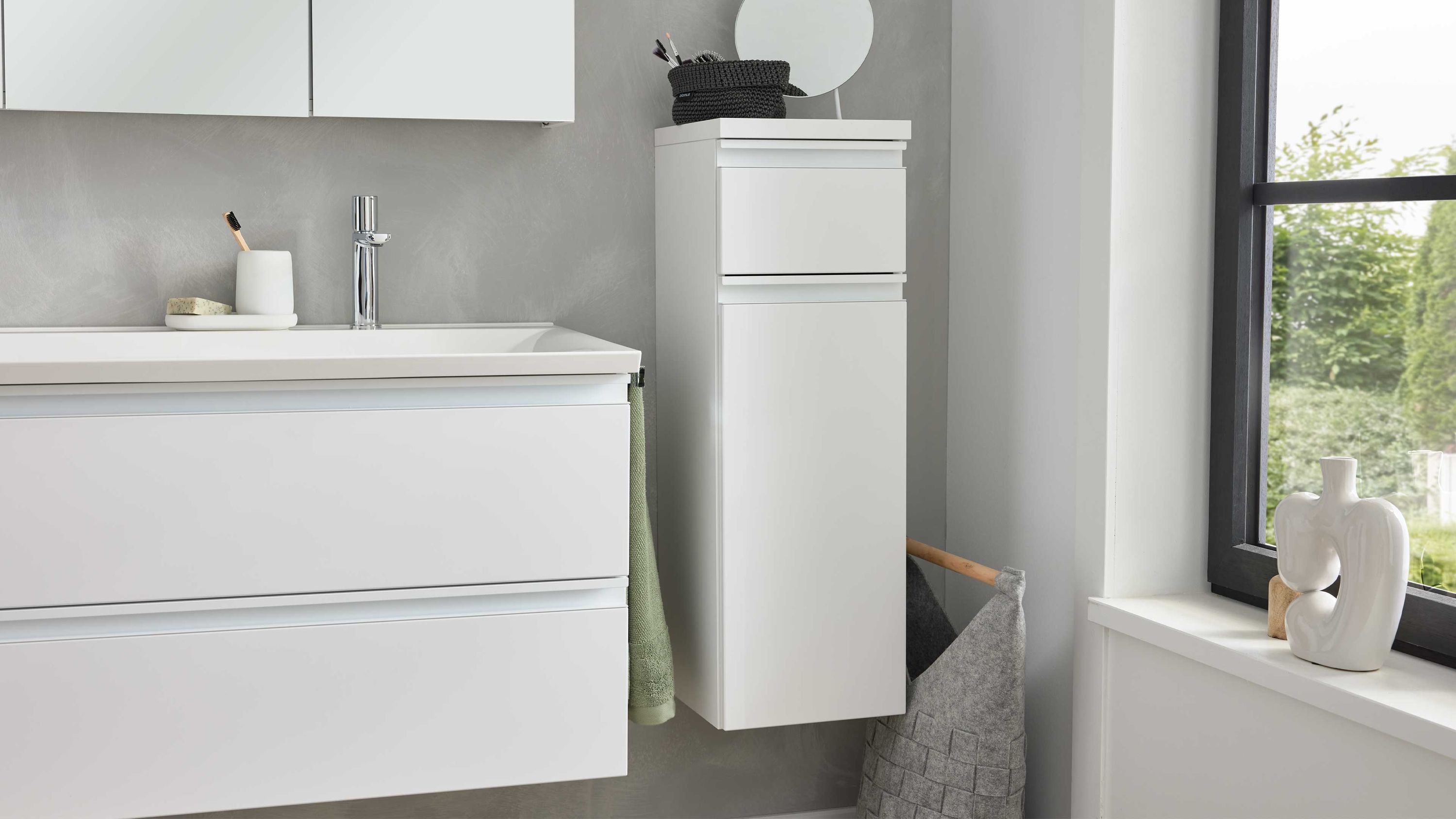 Weißes Highboard mit einer Schublade und einer Tür hängend an grauer Wand im Badezimmer