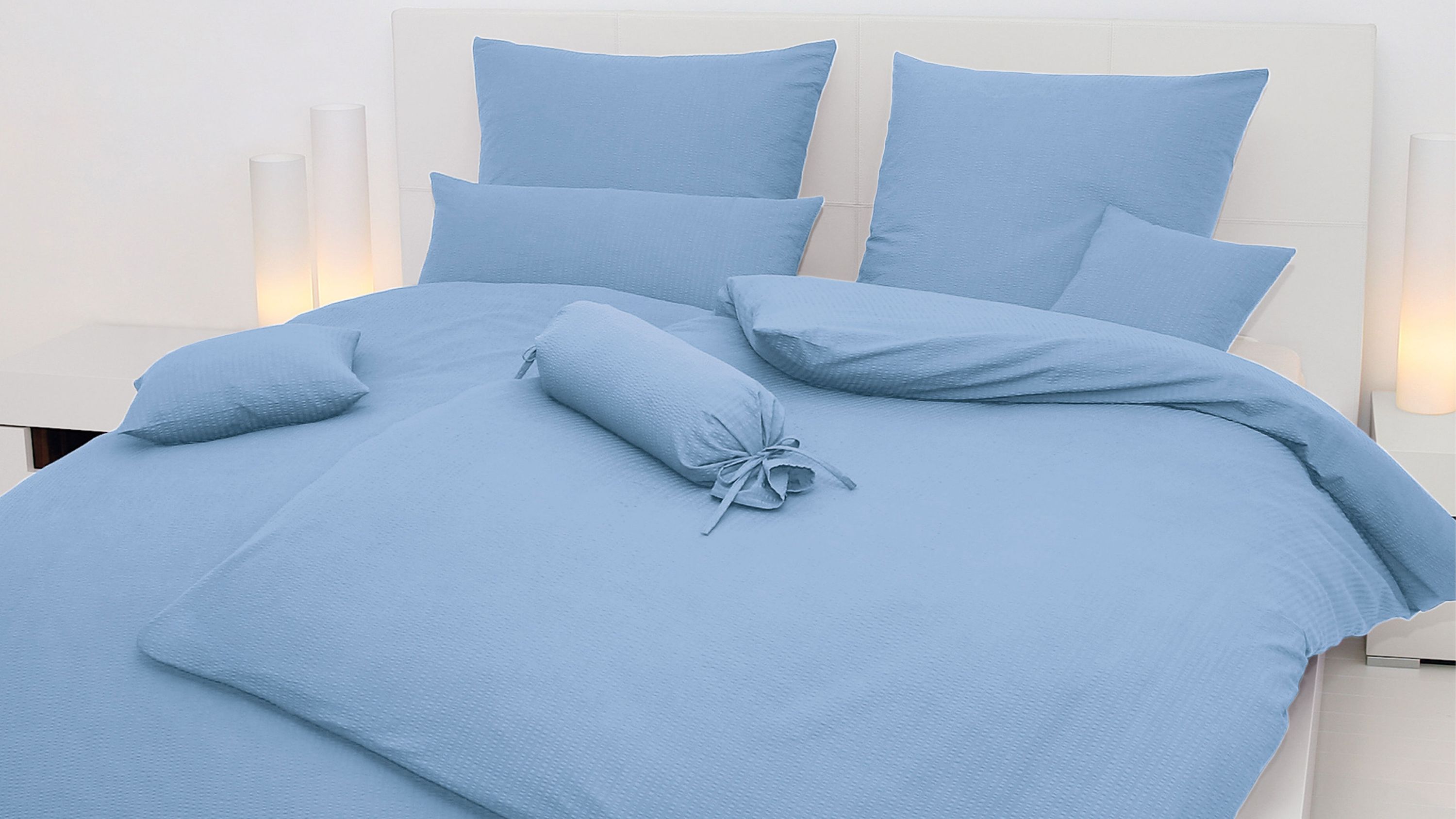 Hellblaue Bettwäsche mit mehreren Kissen und einem runden Kissen auf einem weißen Bett