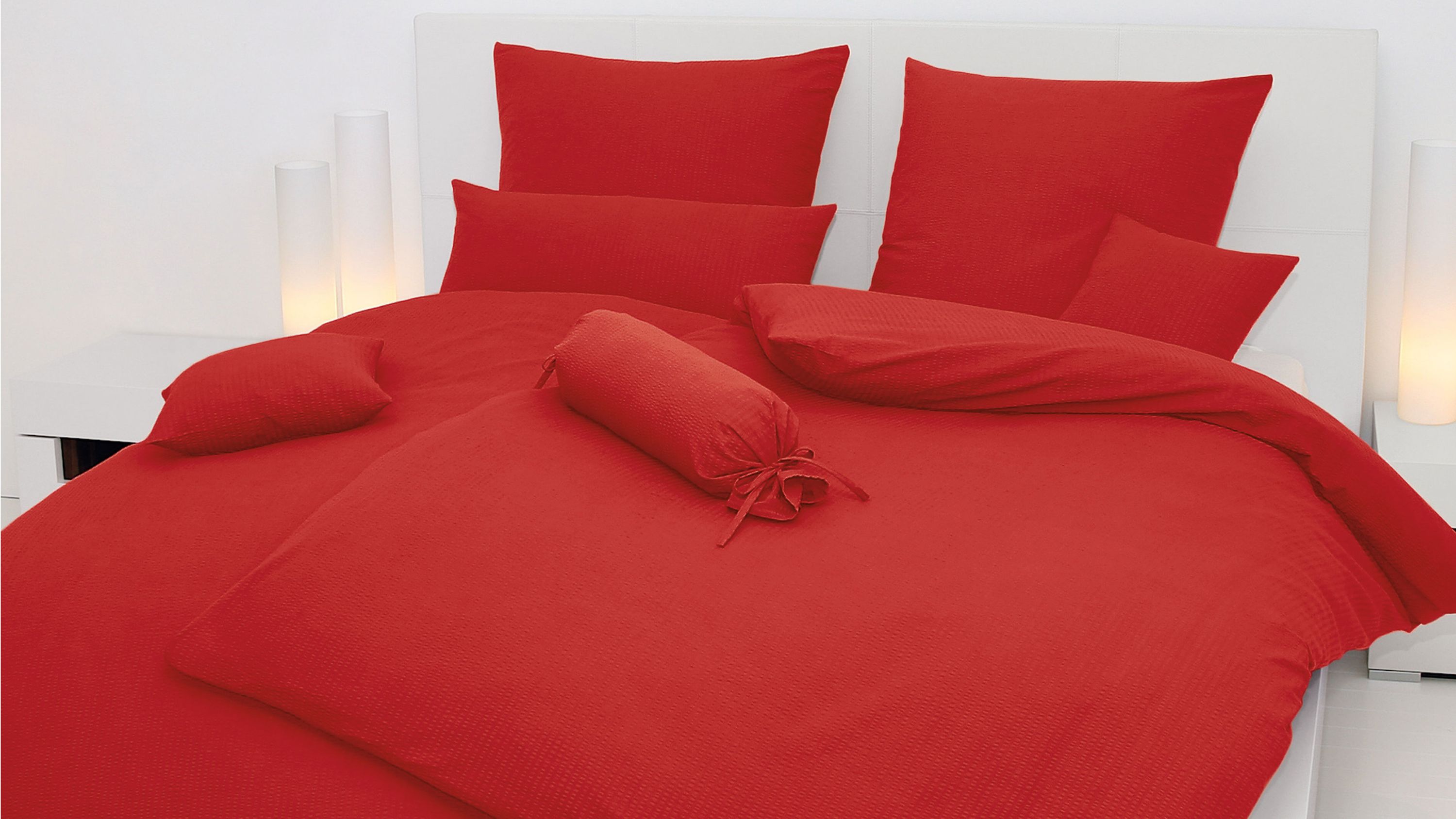 Rotes Bettwäsche-Set mit Kissen und Zierrolle auf weißem Bett mit Kopfteil