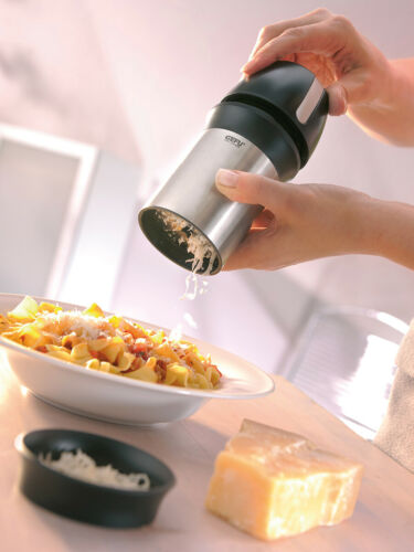 Käsereibe hobelt Käse über einen Teller Pasta  daneben Käseblock und Schale mit Raspeln