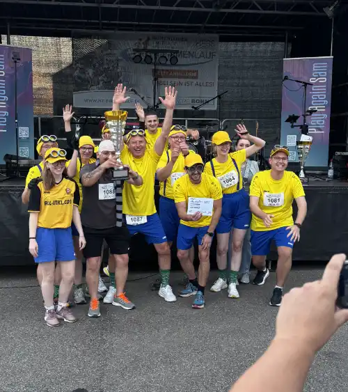 Eine Laufgruppe beim Firmenlauf 2025 feiert vor der Bühne mit einem Pokal für das kreativste Outfit, verkleidet als Minions und Gru.