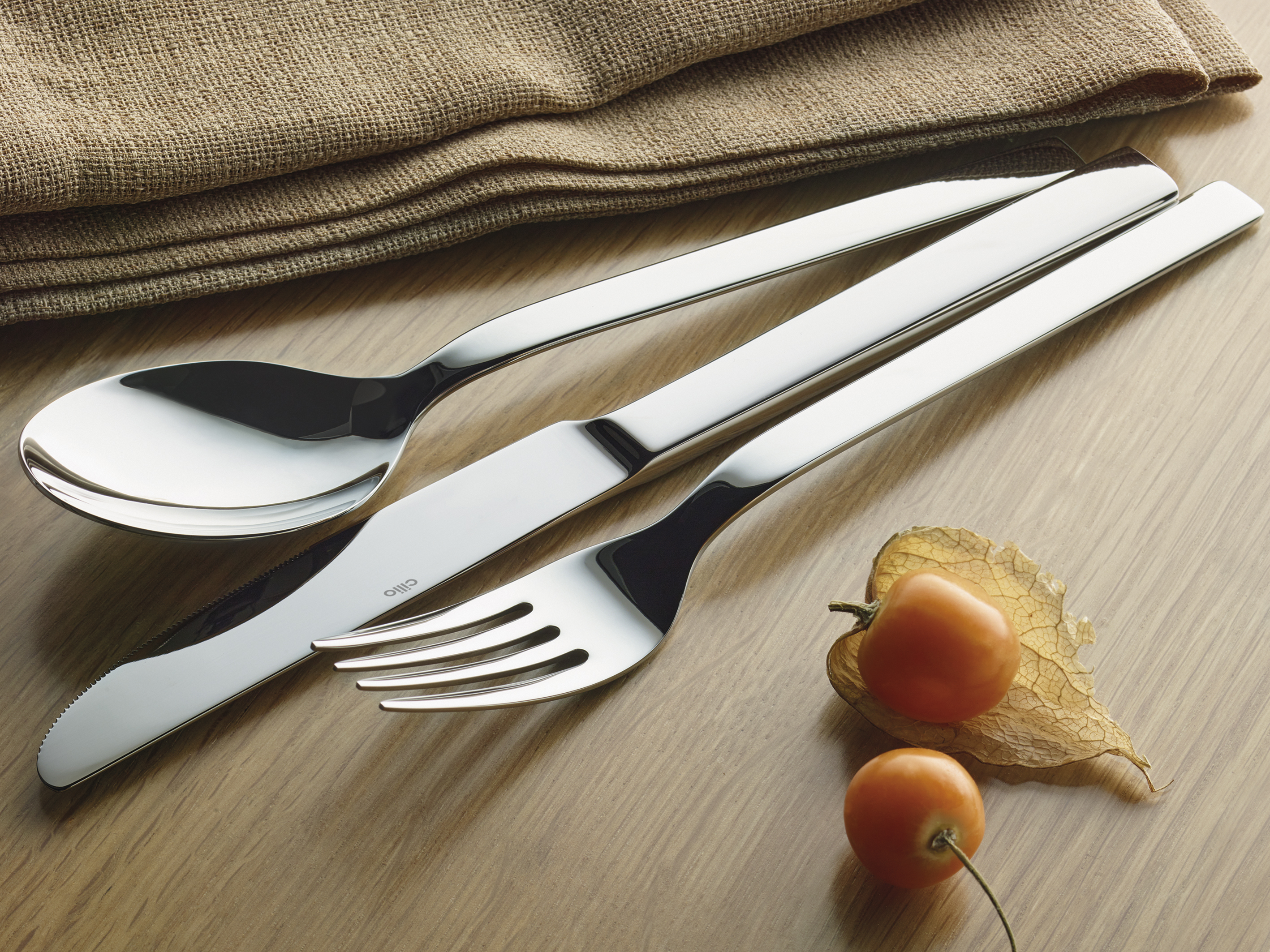 Besteck aus Messer Gabel Löffel aus silberfarbenem Metall auf Holzoberfläche mit braunem Stoff und zwei orangenen Früchten