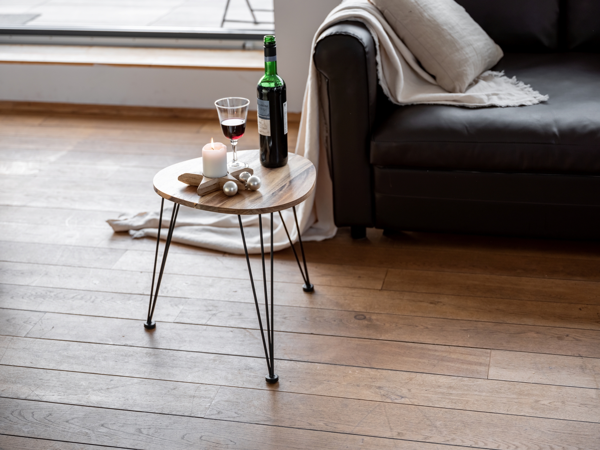 Beistelltisch, stabiles Dreieckdesign Blick aus Sitzhöhe auf runden Holztisch mit Wein und Kerze neben schwarzem Ledersofa im Wohnzimmer.