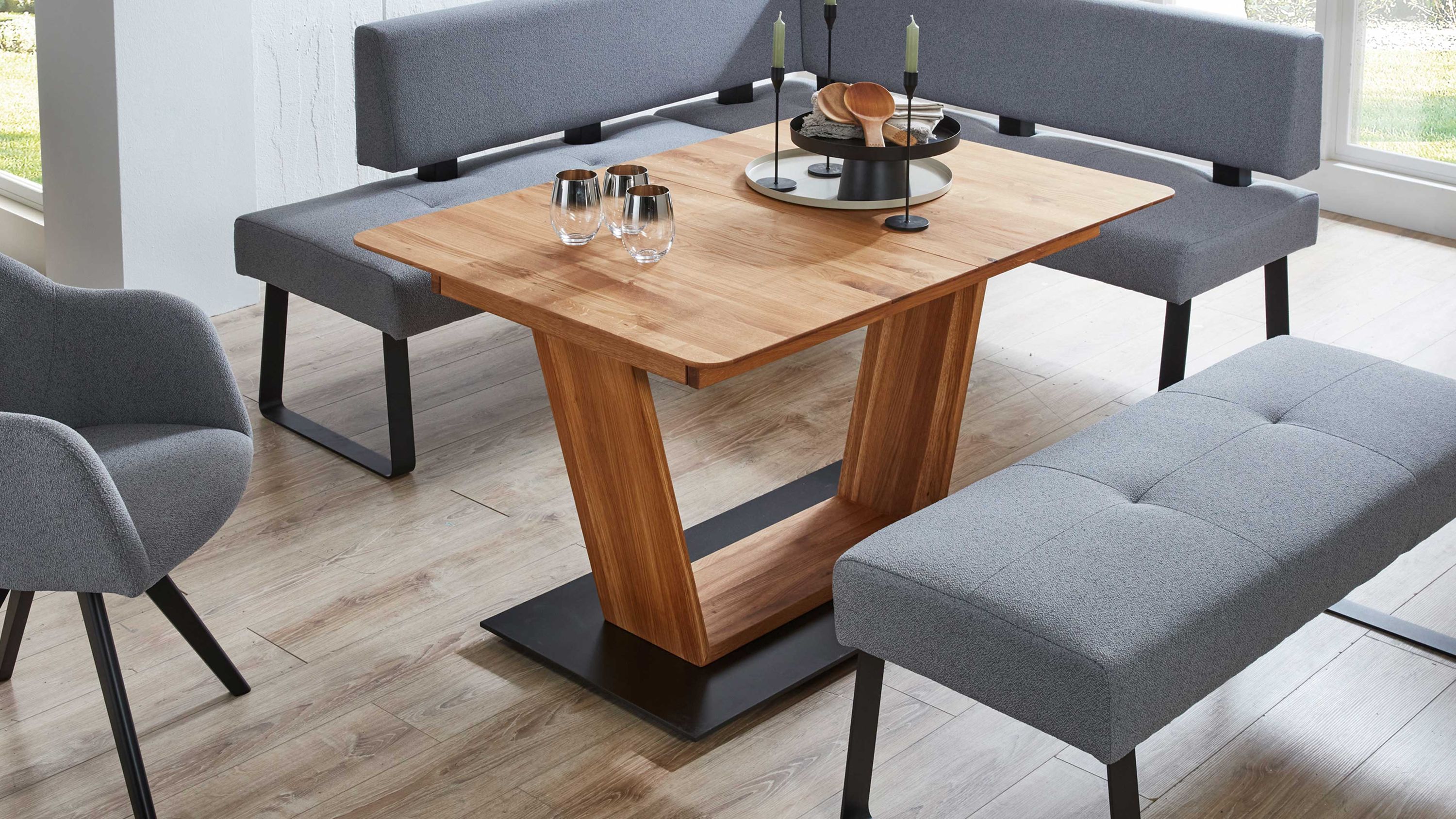 Ausziehtisch mit Holzplatte und V-Gestell auf schwarzer Metallplatte umgeben von grauen Sitzbänken und Stuhl.