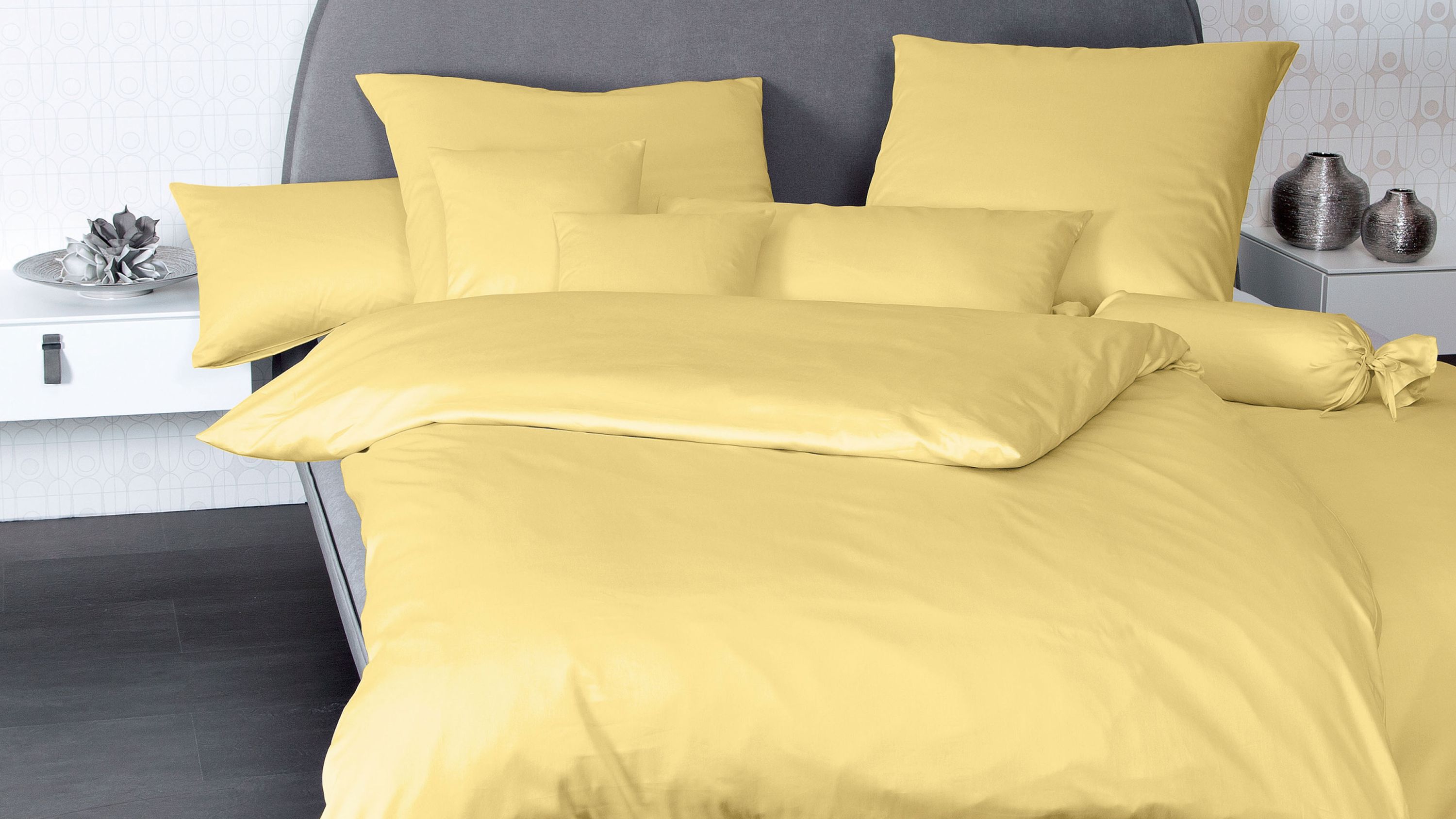 Gelbe Bettwäsche mit mehreren Kissen auf einem grauen Bett mit weißem Nachttisch
