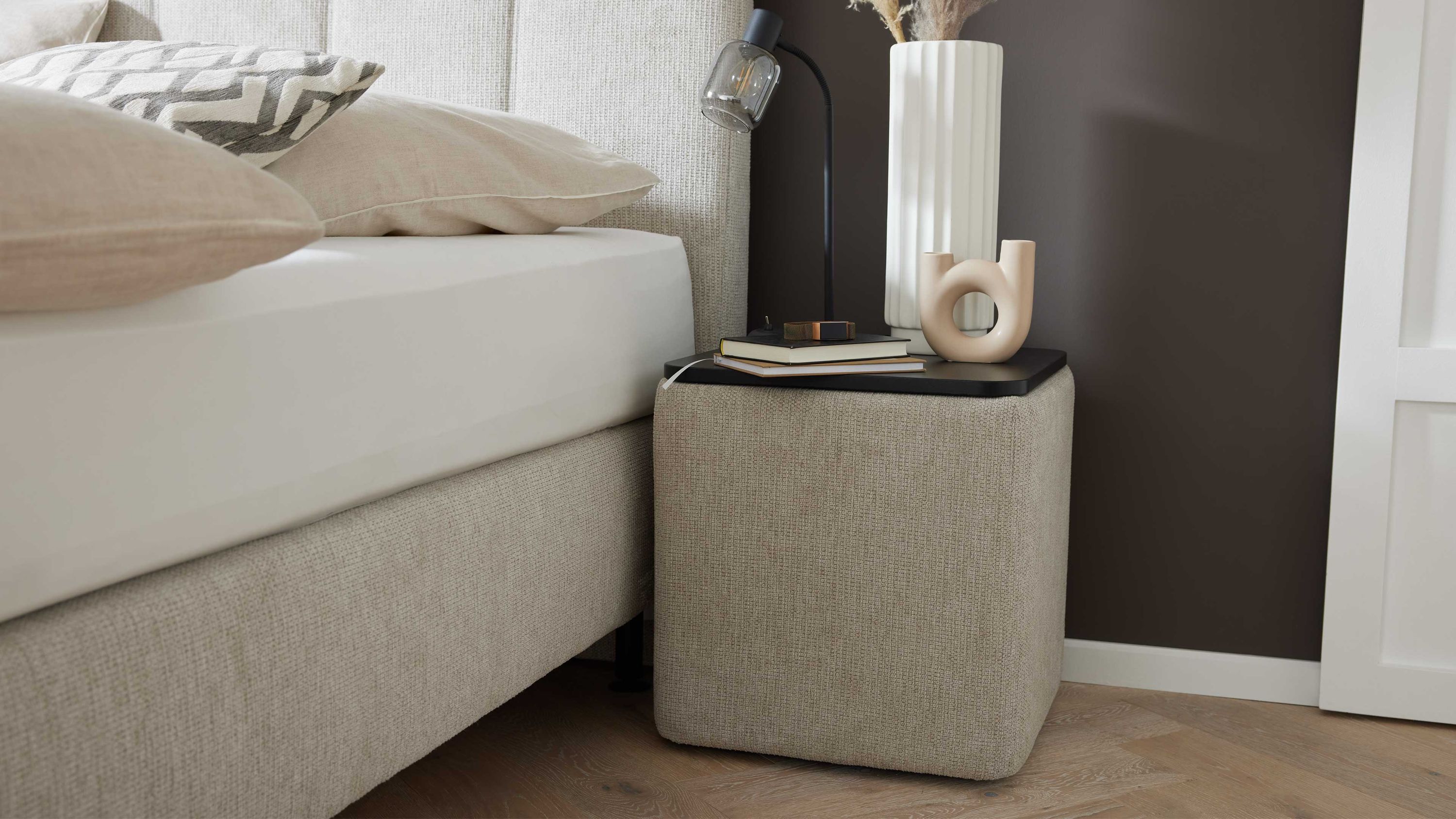 Kubische Nachtkonsole mit Stoffbezug neben Boxspringbett Kubische Nachtkonsole mit naturfarbenem Stoffbezug und schwarzer Ablagefläche neben einem hellen Boxspringbett