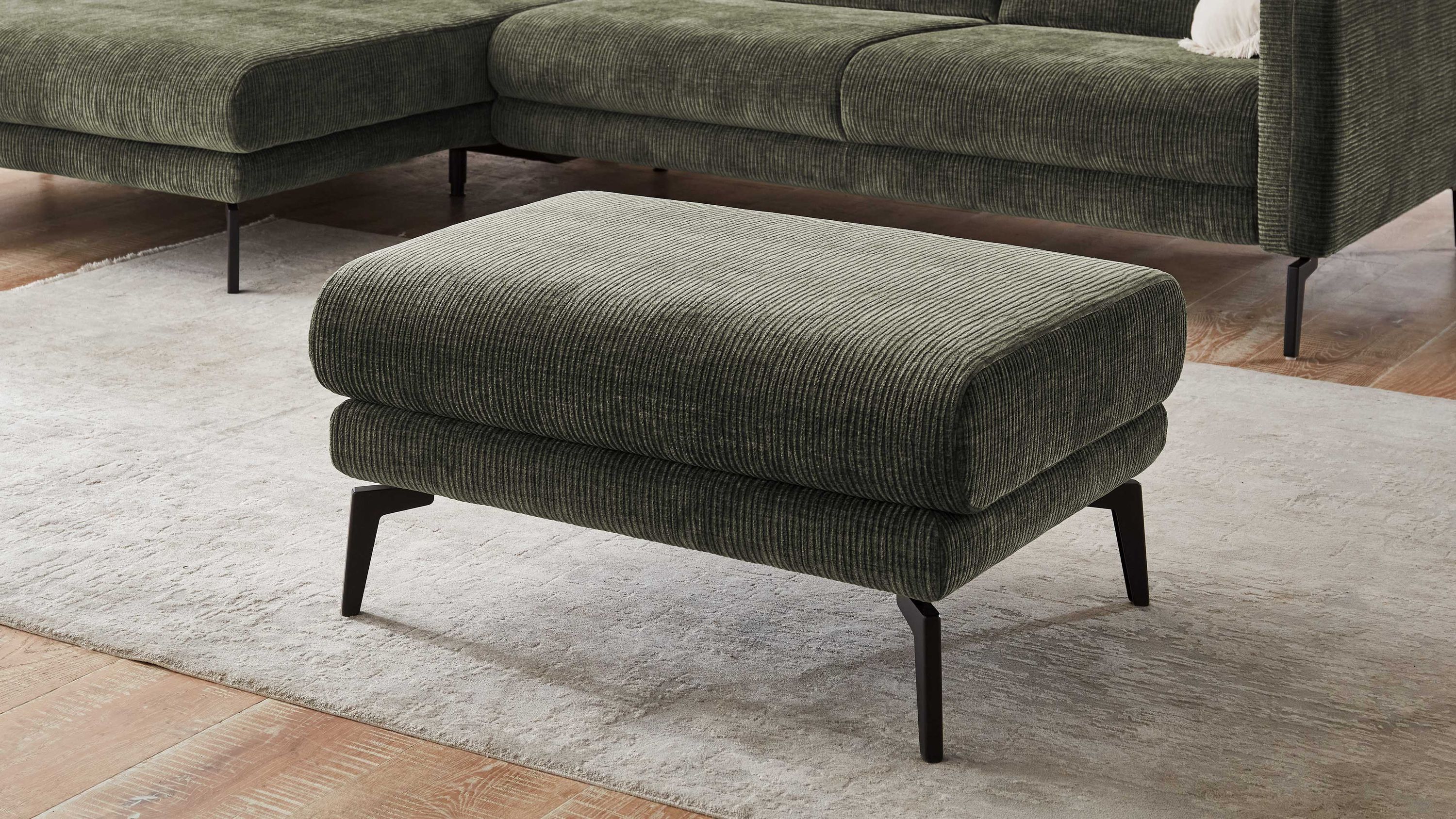 Rechteckiger Hocker mit olivgrünem Flachgewebe und schwarzen Metallfüßen vor grünem Sofa