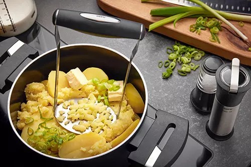 Kartoffelstampfer mit schwarzem Griff in Topf mit gekochten Kartoffeln auf heller Fläche