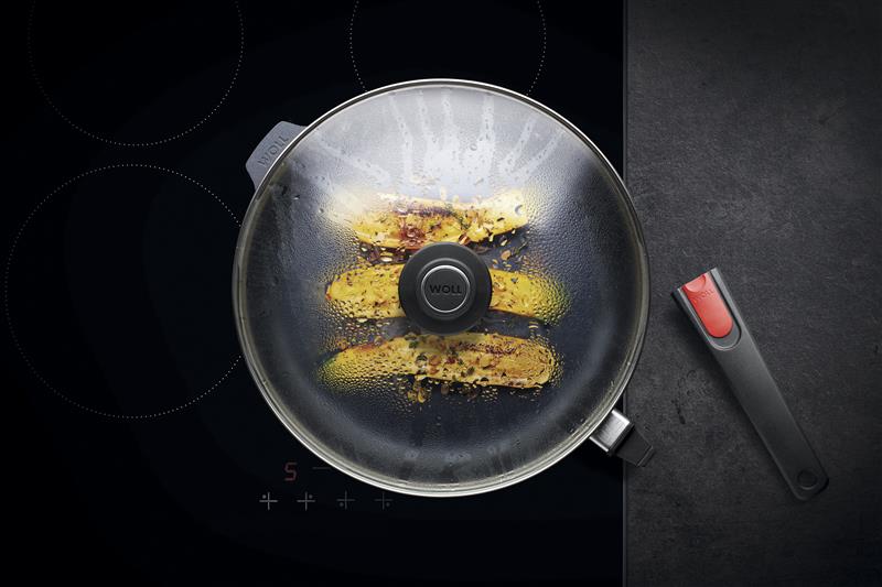 Pfanne mit Glasdeckel auf Kochfeld, darin gelbe Speisen mit Gewürzen, Griff liegt daneben.