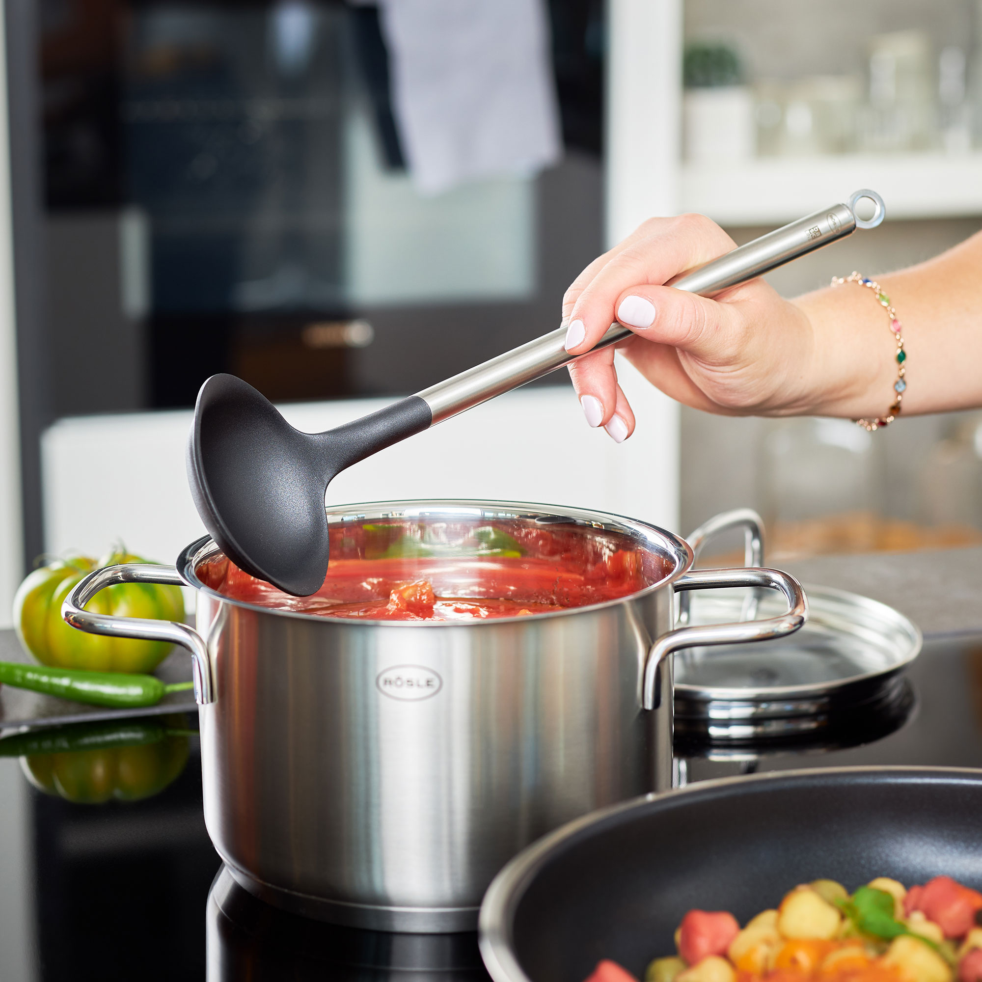 Schöpflöffel aus Edelstahl rührt tomatenbasiertes Gericht im Topf seitlich links aufgenommen mit Paprika im Hintergrund