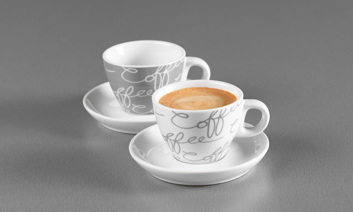 Zwei Espressotassen mit Untertassen in Weiß und Grau mit Schriftzug auf grauem Untergrund