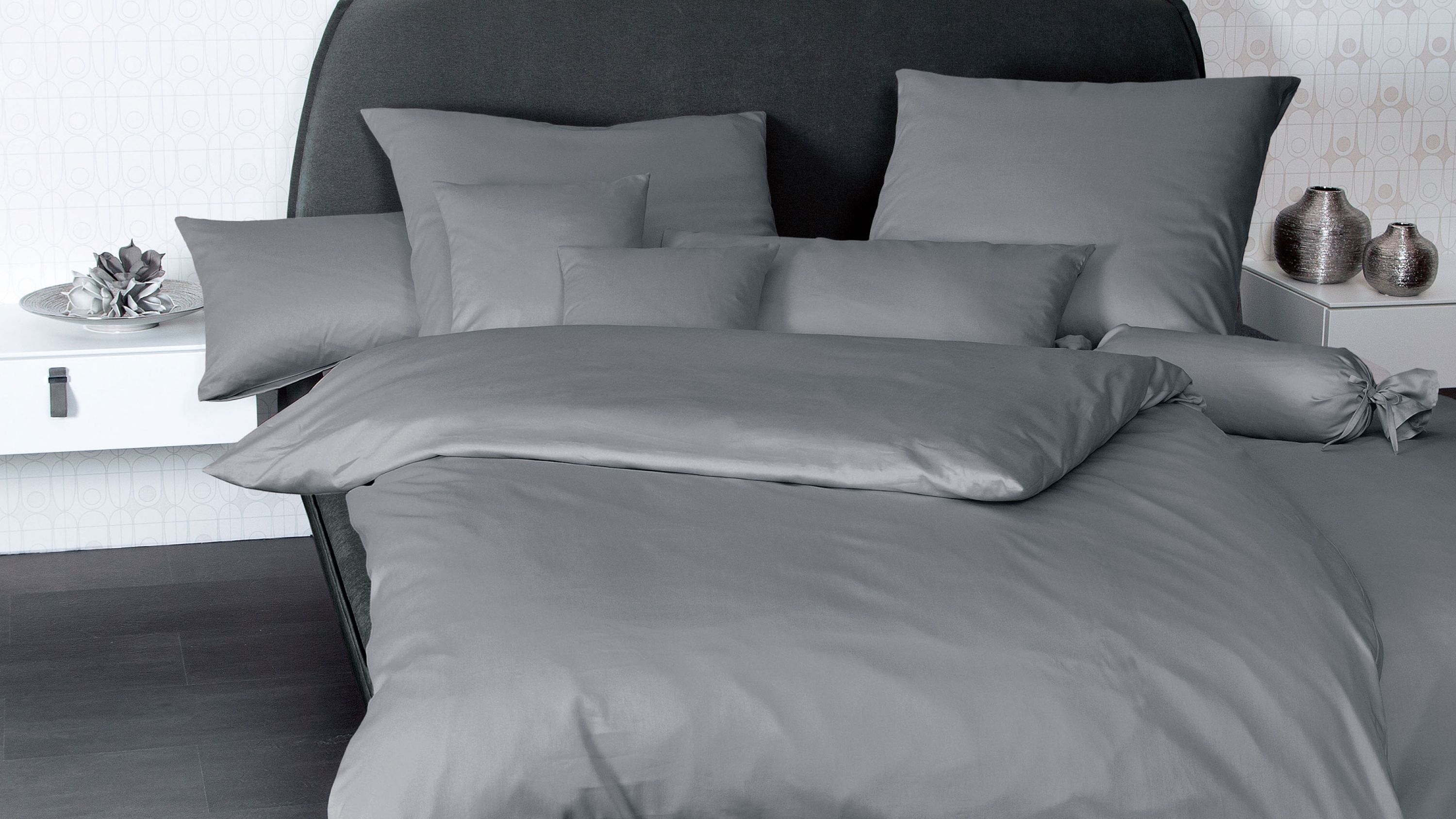 Bett mit grauer Bettwäsche bestehend aus Kissen und Decke vor dunklem Kopfteil