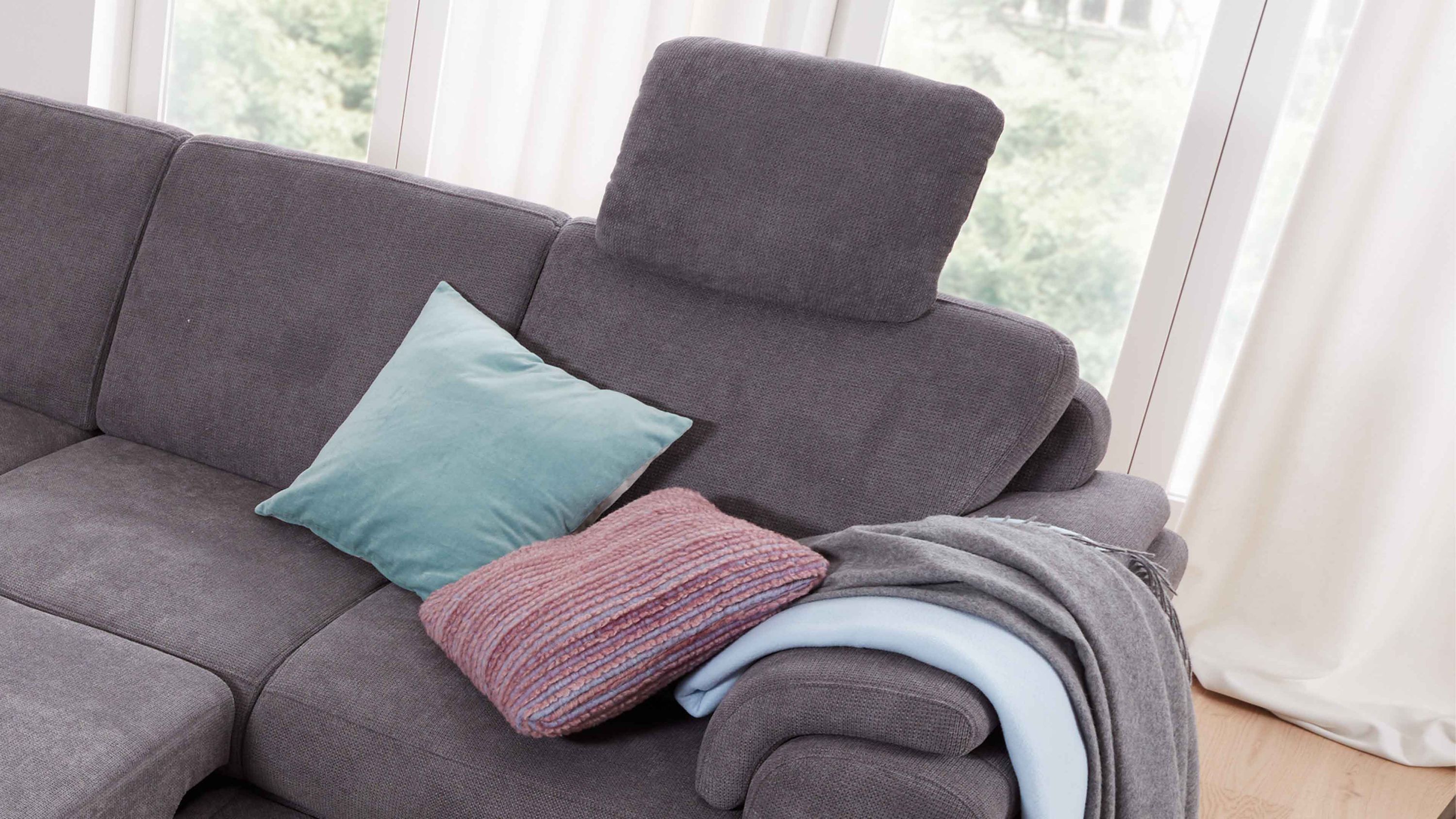 Graues Sofa mit verstellbarer Kopfstütze und zwei Kissen sowie einer Decke auf der Armlehne