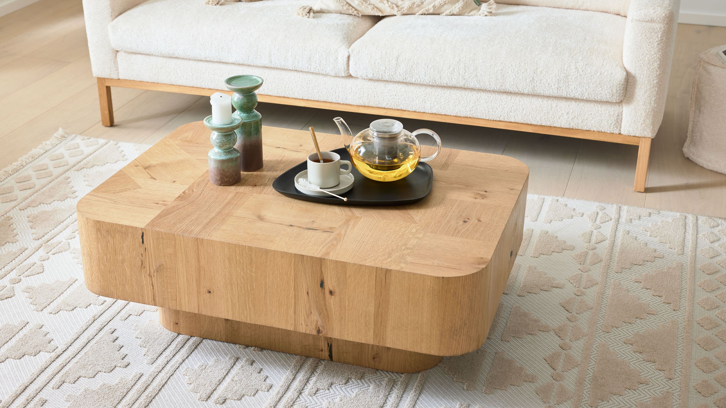 Rechteckiger Couchtisch mit abgerundeten Ecken aus hellem Holz mit Teekanne Tasse und Kerzen auf Teppich vor weißem Sofa