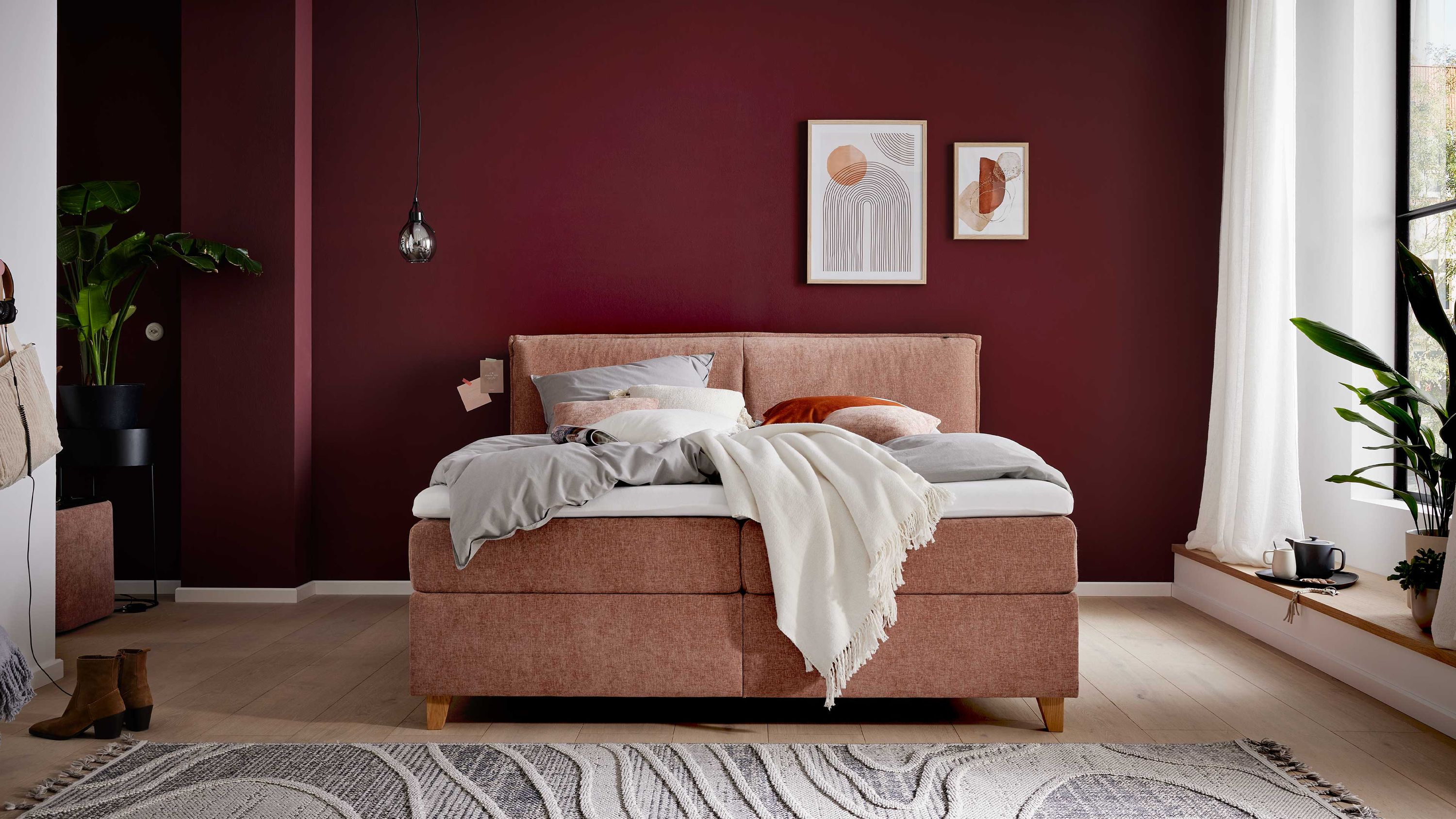 Boxspringbett mit sienafarbenem Stoffbezug und hellen Holzfüßen vor dunkelroter Wand mit zwei Bildern