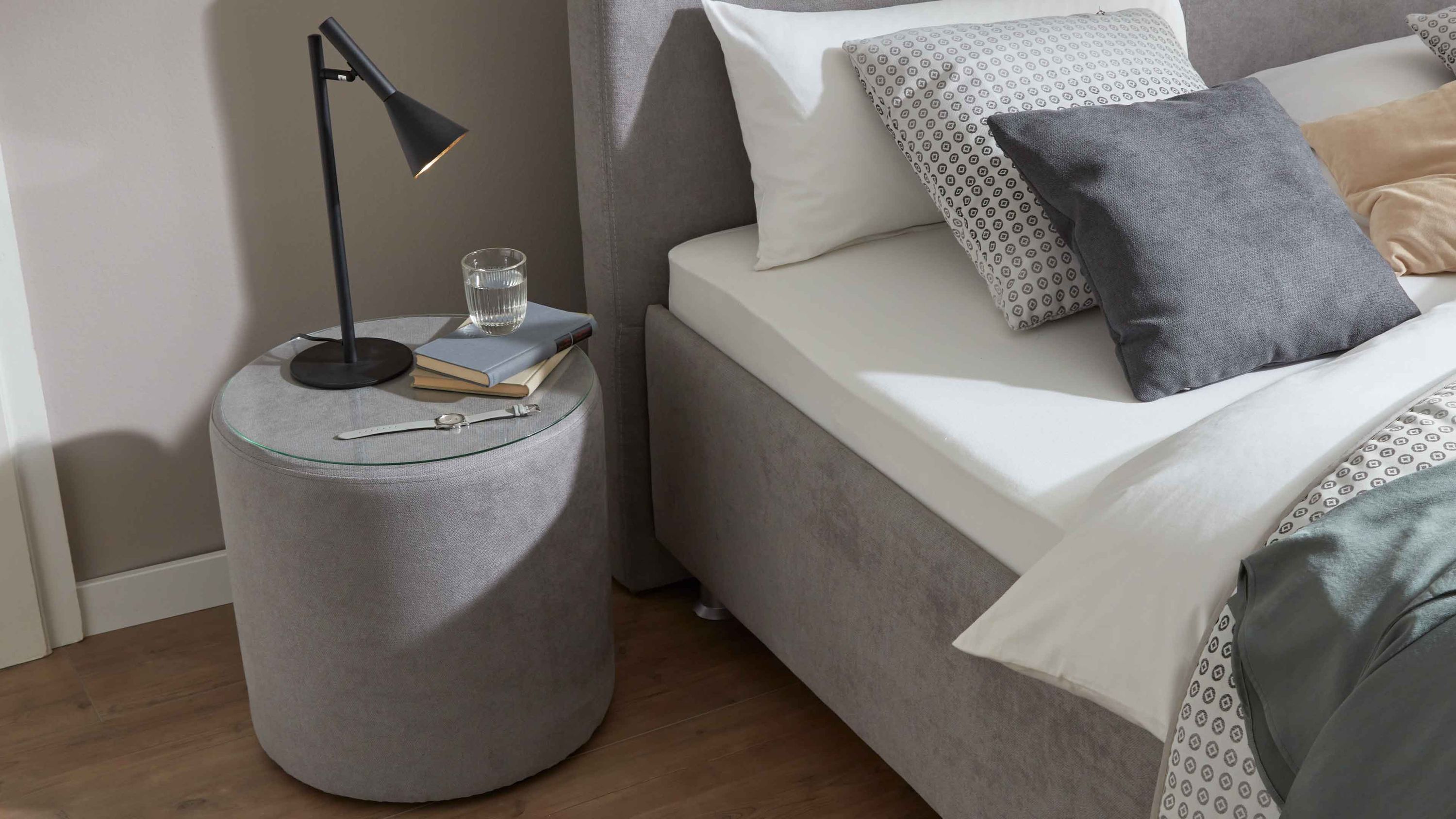 Runde graue Nachtkonsole mit Glasdeckel neben Bett Runde graue Nachtkonsole mit klarem Glasdeckel neben einem grauen Bett mit weißen und gemusterten Kissen