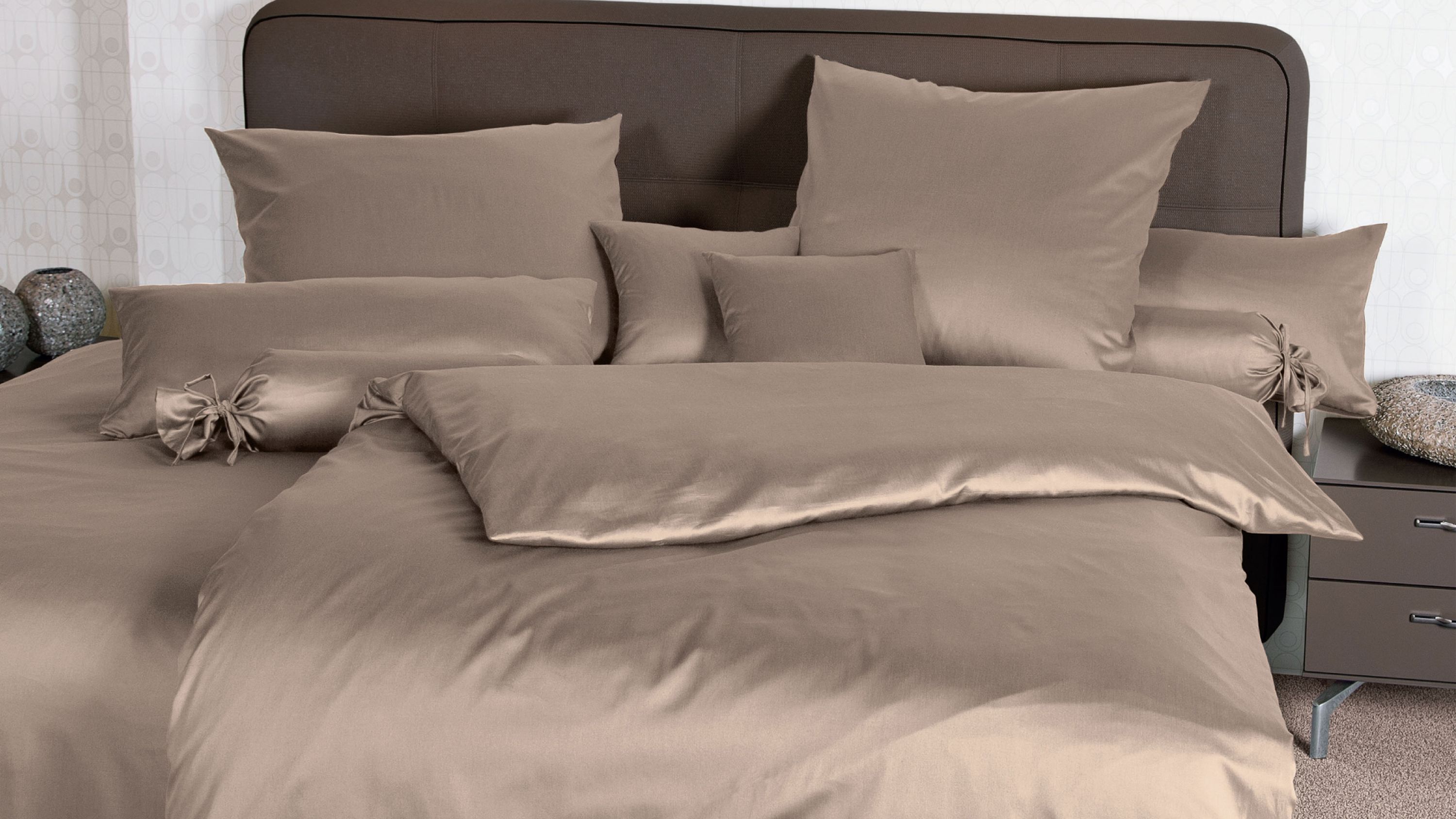 Bett mit taupefarbener Bettwäsche bestehend aus Kissen und Decke vor braunem Kopfteil