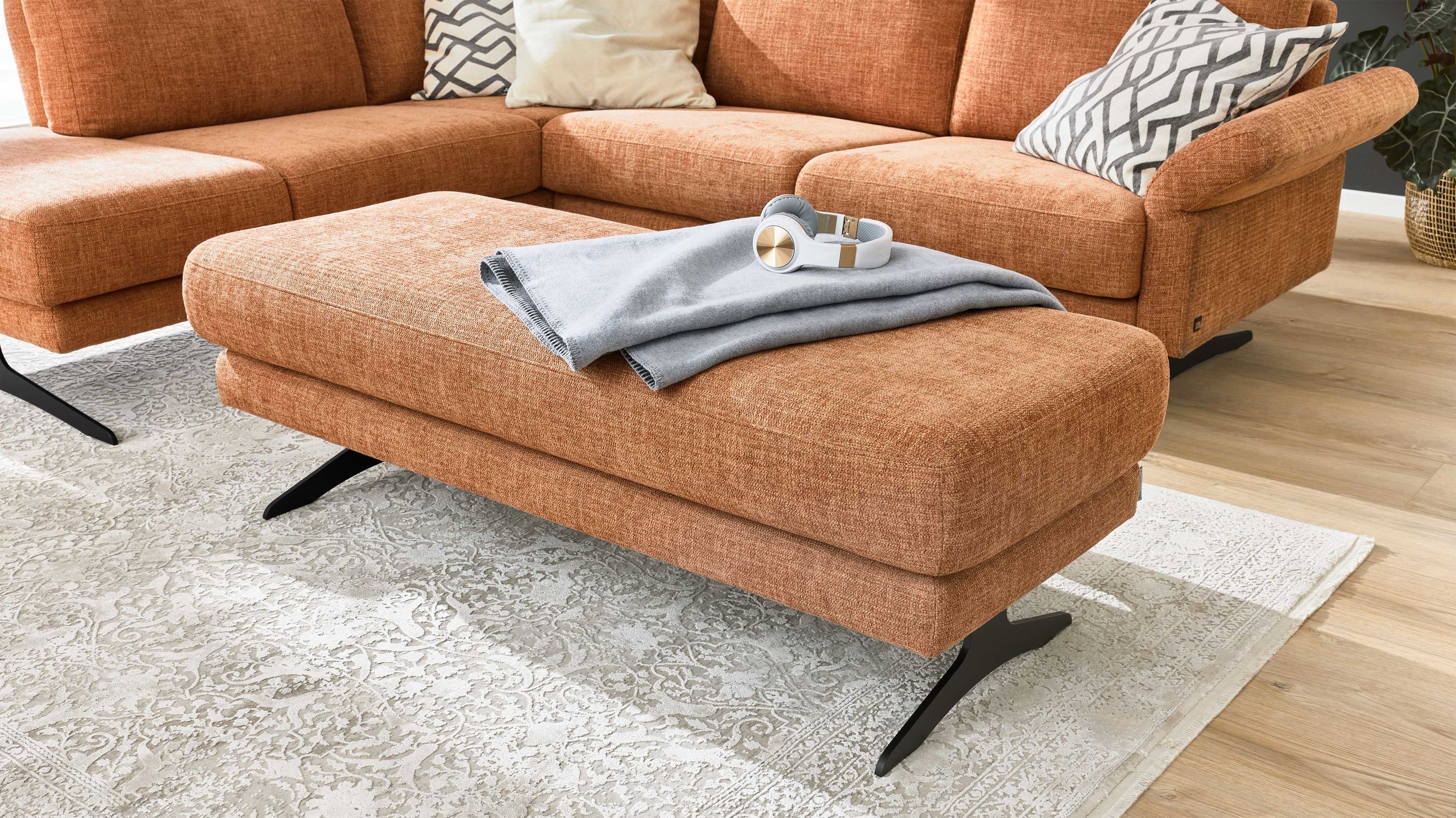 Rechteckiger Hocker in puderfarbenem Stoff mit schwarzen Metallfüßen vor einem Sofa mit gleichen Stoffbezug