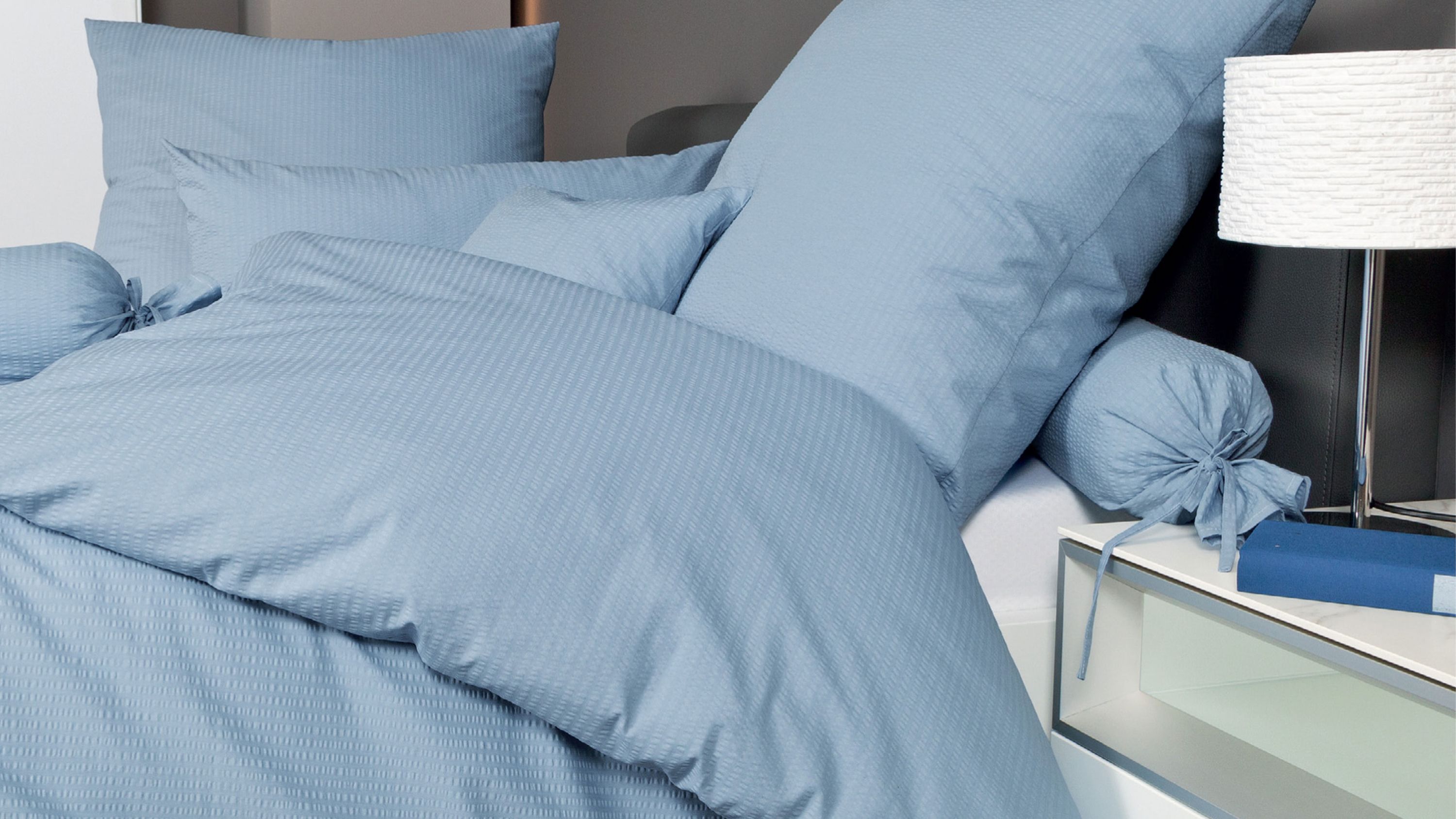 Bettwäsche-Set in hellblau mit Kissen und Zierkissen auf einem Bett mit Nachttisch und Lampe