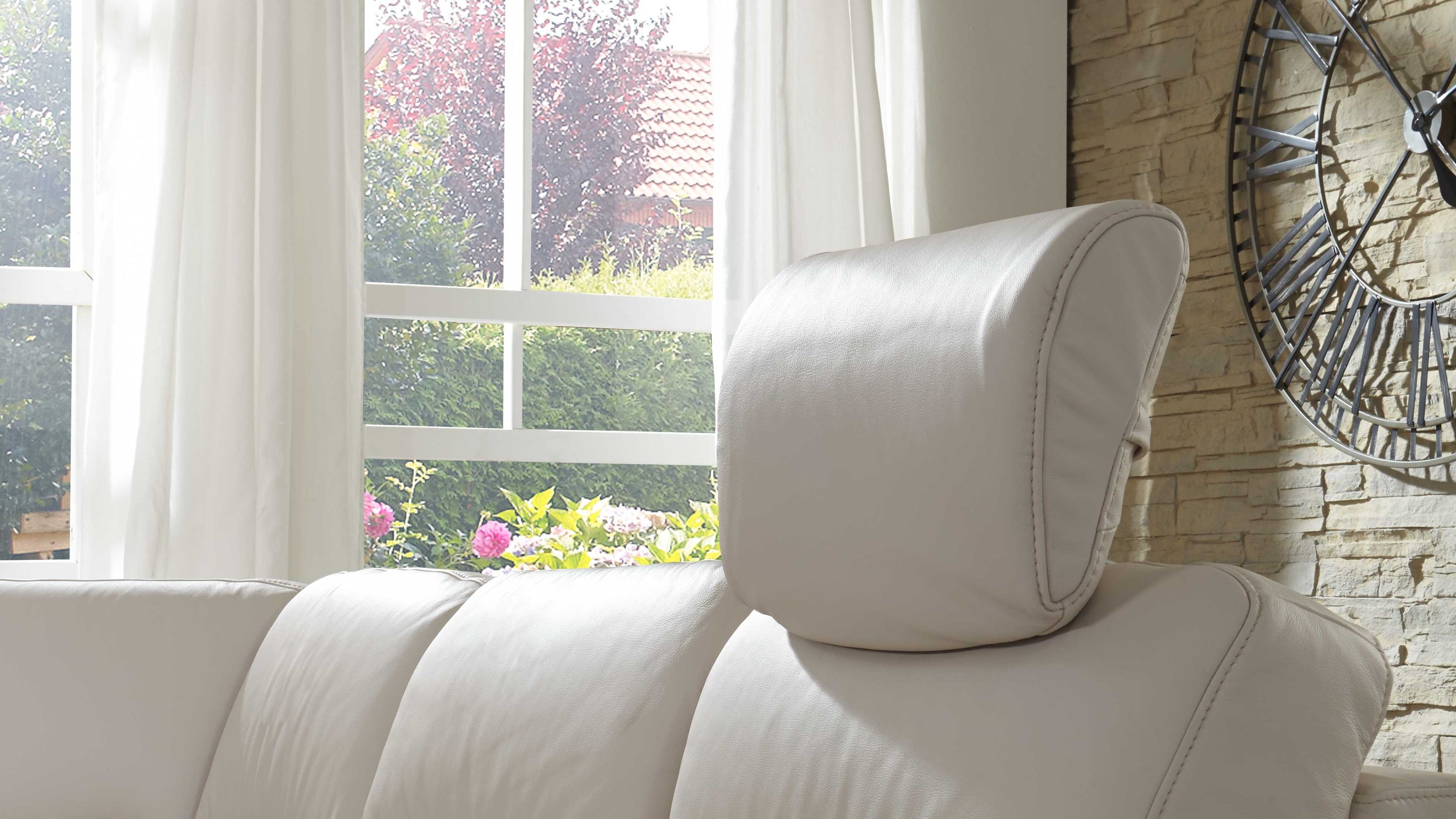 Beige Kopfstütze auf einem hellen Ledersofa vor Fenster mit weißen Vorhängen und Steinwand mit großer Uhr