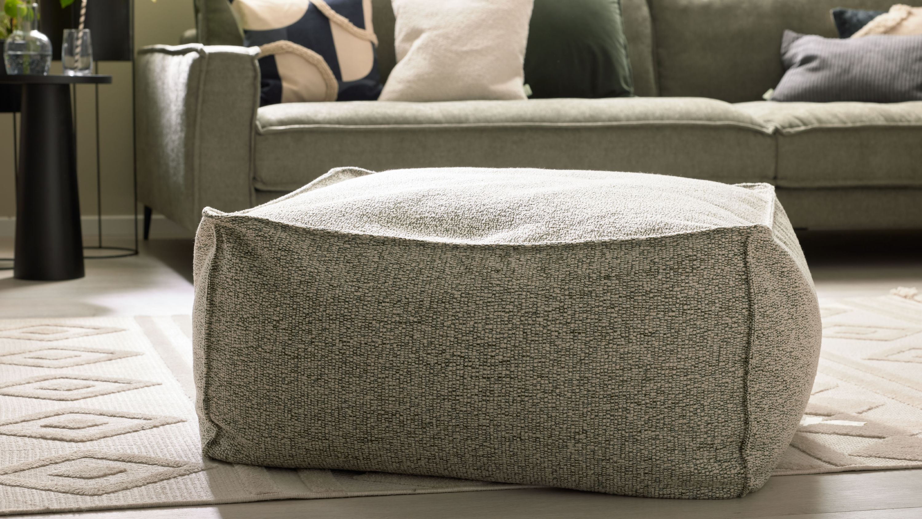 Quadratischer Hocker mit meliertem stoffbezug in schildkrötenfarben auf hellem teppich vor grünem sofa