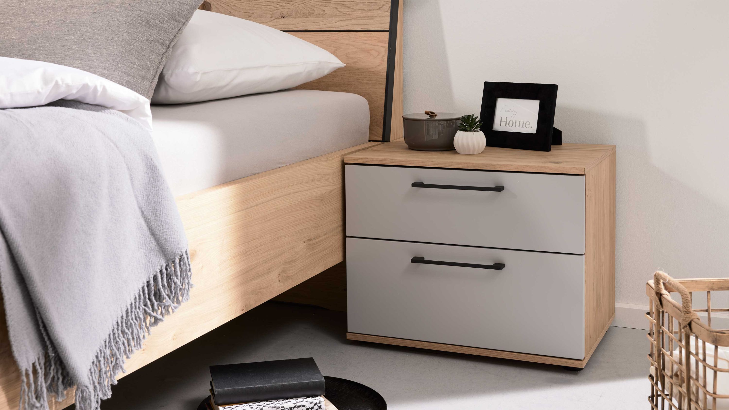 Nachtkommode mit zwei taupefarbenen Schubladen und Rahmen in heller Holzoptik neben einem Bett