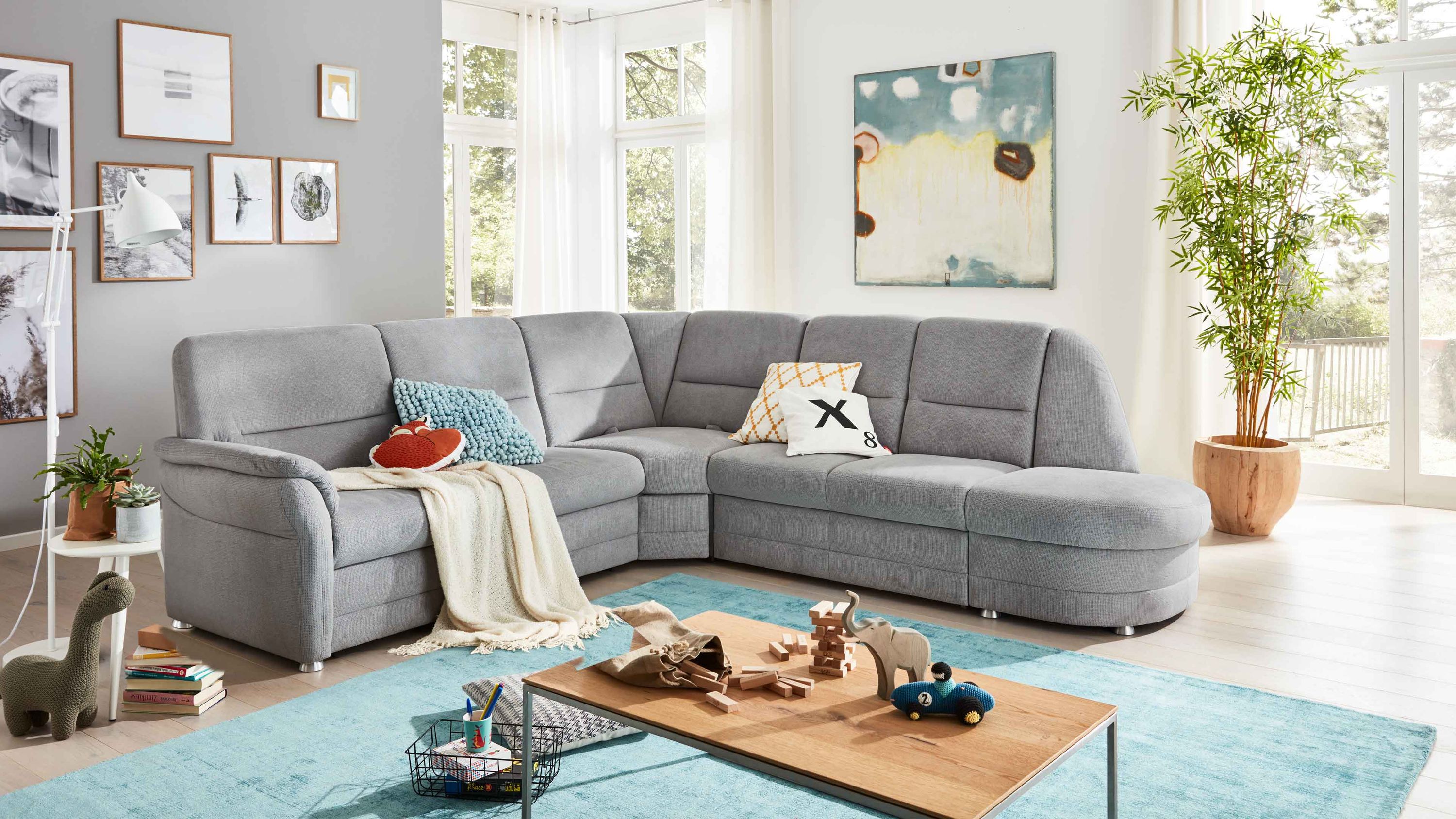 Ecksofa mit silberfarbenem Stoffbezug und Metallfüßen in hellem Wohnzimmer mit Teppich und Dekokissen