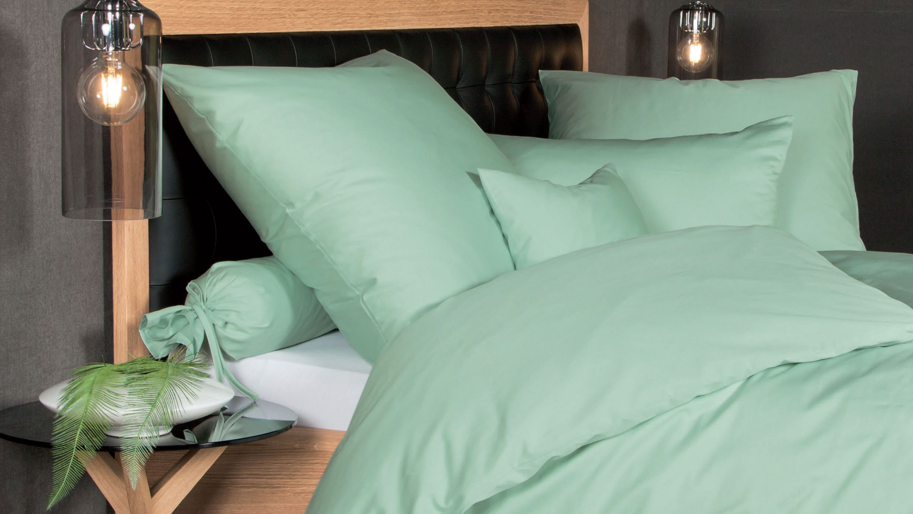 Bett mit Kopfteil aus Holz und schwarzem Polster jadegrüne Bettwäsche mit mehreren Kissen und Zierrolle