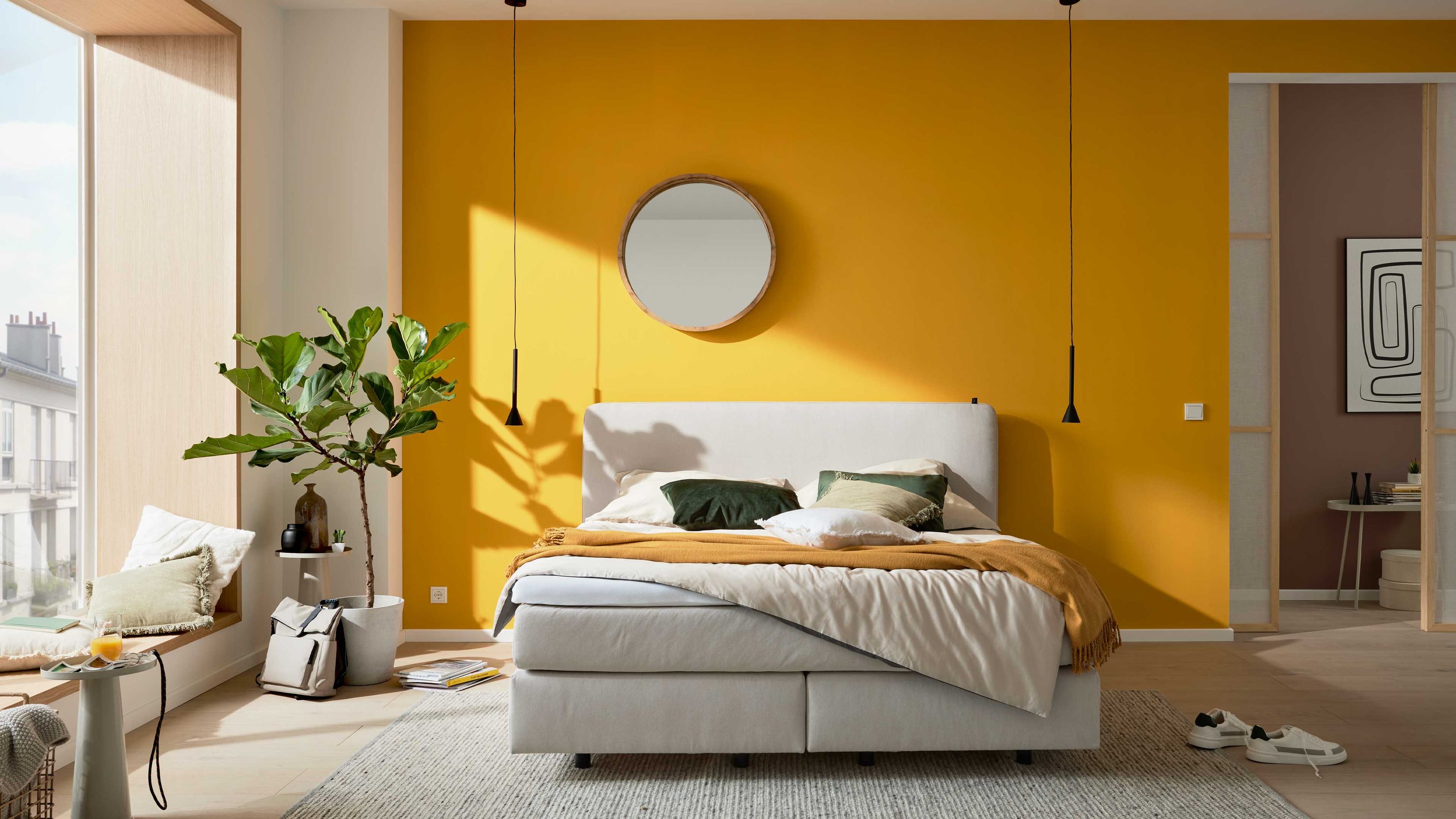 Boxspringbett mit naturfarbenem Stoffbezug und gepolstertem Kopfteil vor gelber Wand mit rundem Spiegel