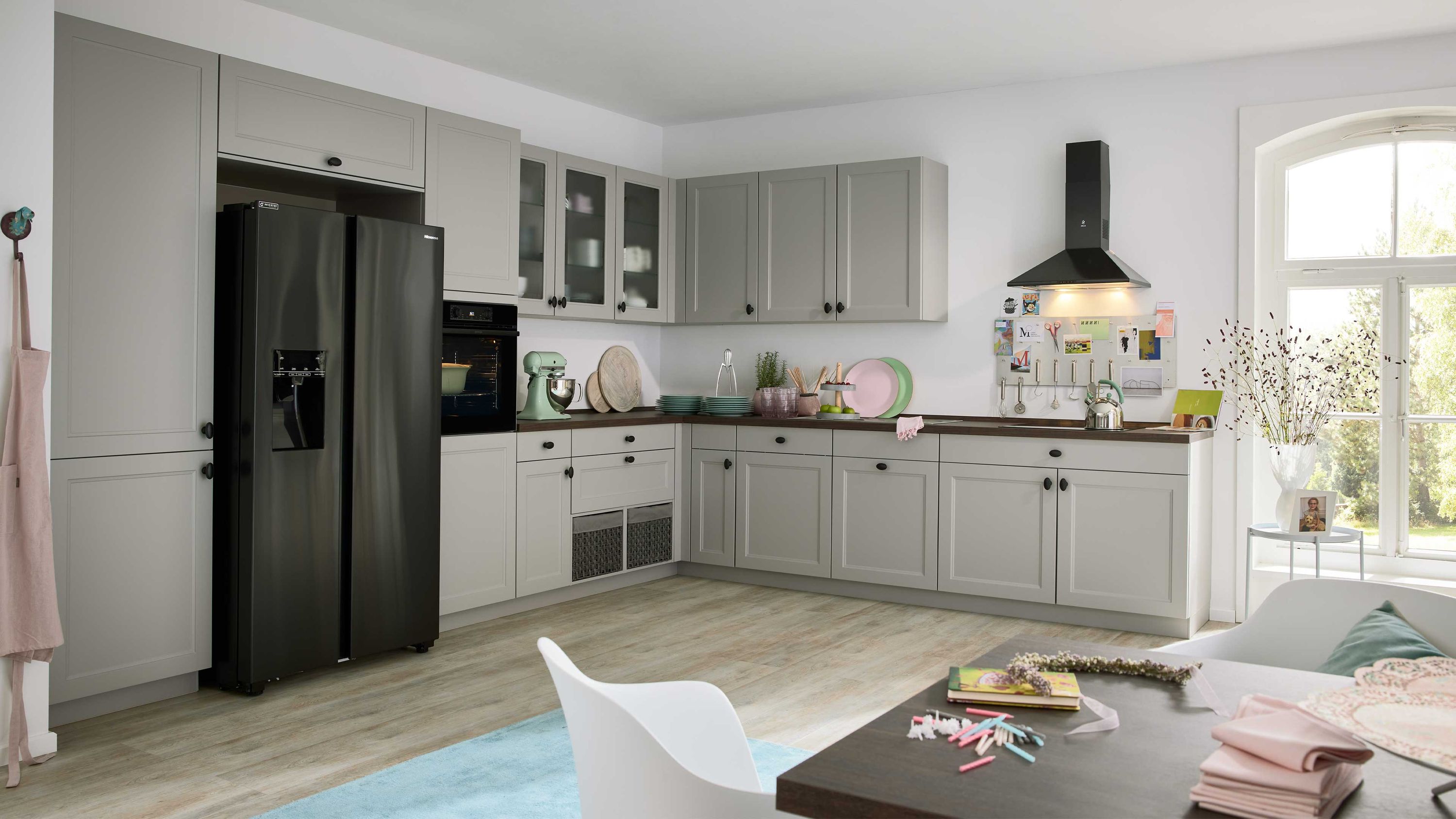 Moderne Küche mit grauen Fronten und schwarzer Ausstattung Küche mit grauen Schränken schwarzem Kühlschrank Holzfußboden Esstisch mit Büchern Tageslicht durch Seitenfenster