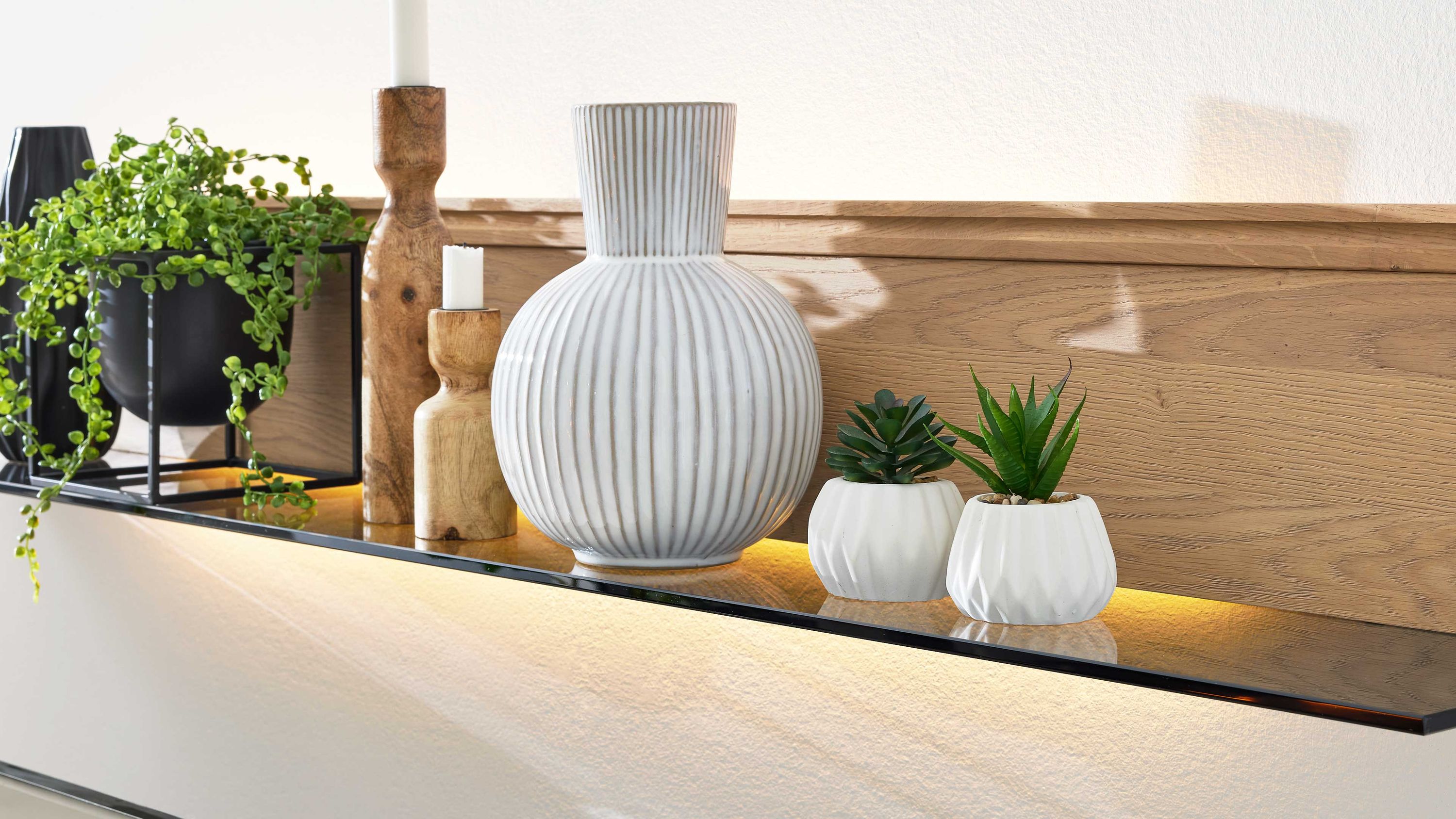 Wandregal mit LED-Profilbeleuchtung und Dekoration Schwarzes Wandregal mit LED-Profilbeleuchtung und Dekoration aus weißen Vasen Holzkerzenhaltern und grünen Pflanzen
