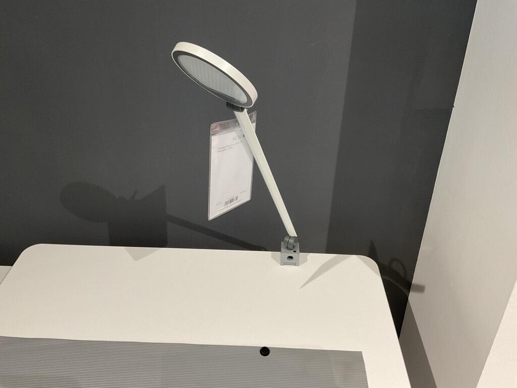 Weiße Tischleuchte mit rundem Kopf und geradem Arm, seitlich an einer Tischkante befestigt.