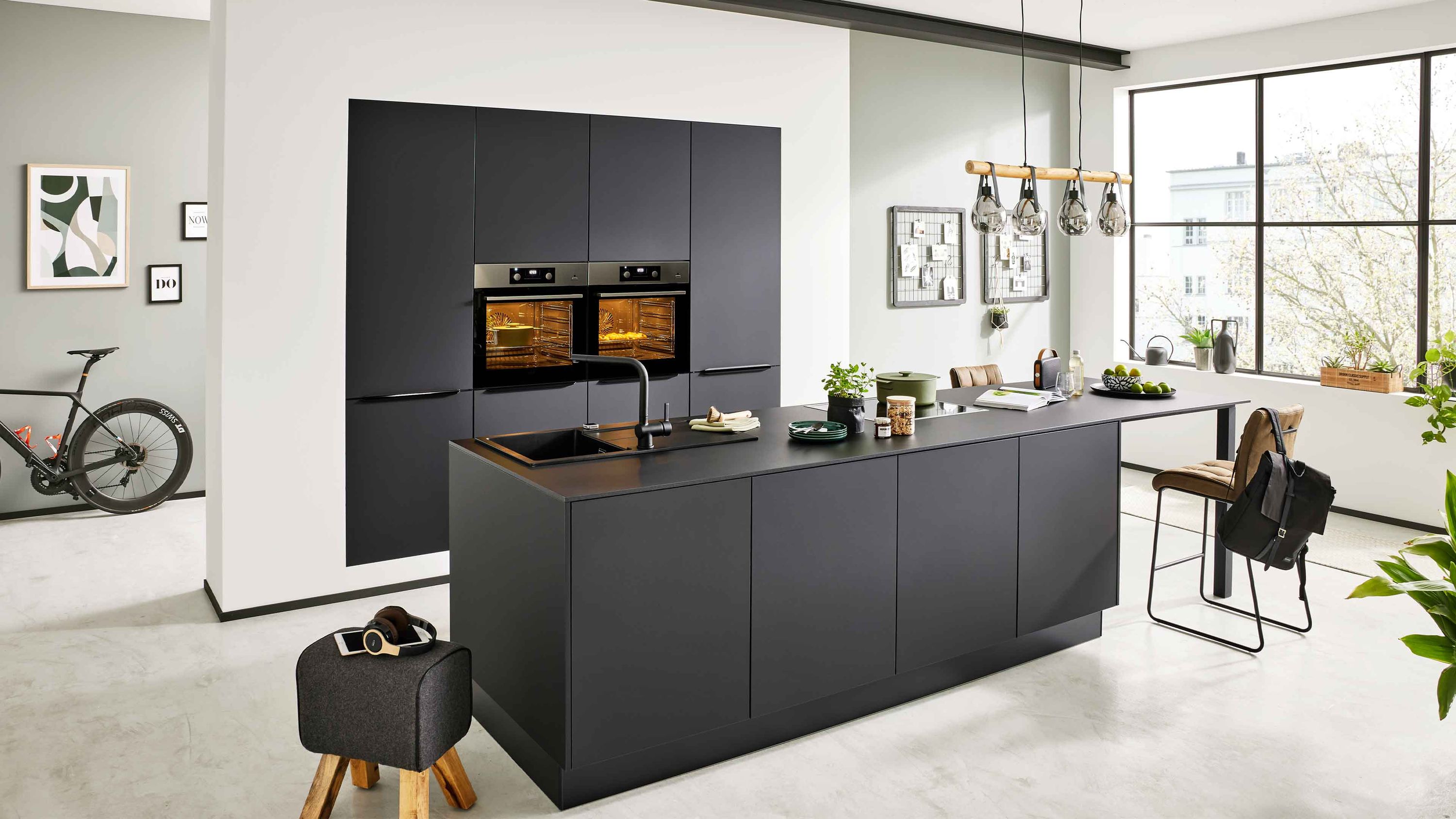 Küche mit dunkler Insel und modernem Loft-Flair Dunkle Schränke schwarze Insel mit Barhockern Hängelampen Fensterlicht Pflanzen Fahrrad im Hintergrund