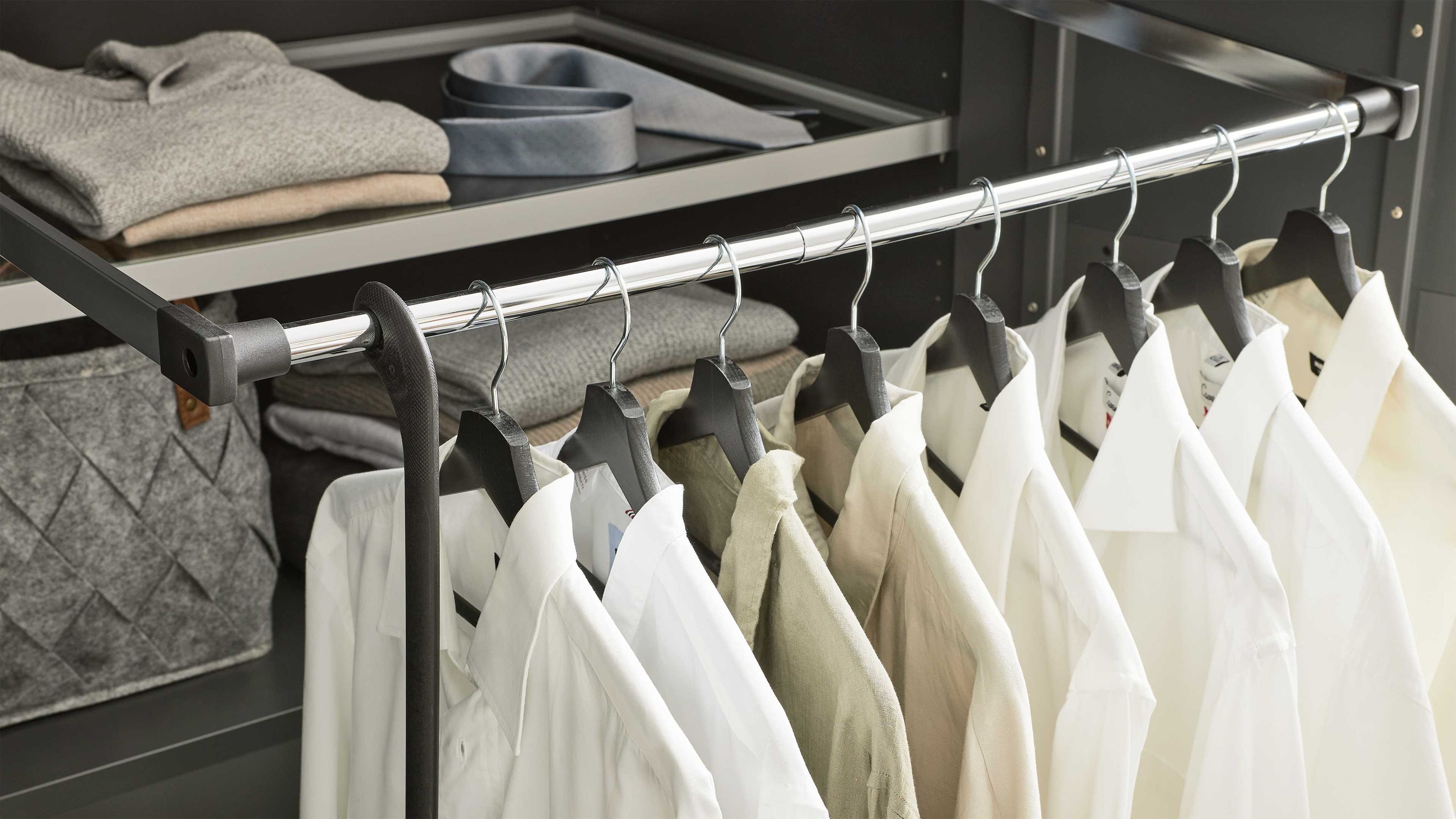 Verchromte Kleiderstange mit schwarzen Kleiderbügeln und weißen sowie beigen Hemden in einem schwarzen Schrank