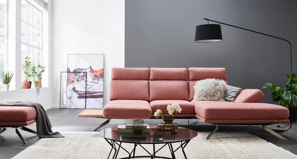 Modernes Wohnzimmer mit einem roséfarbenen Ecksofa, einem kleinen Teppich und Pflanzen im Hintergrund.