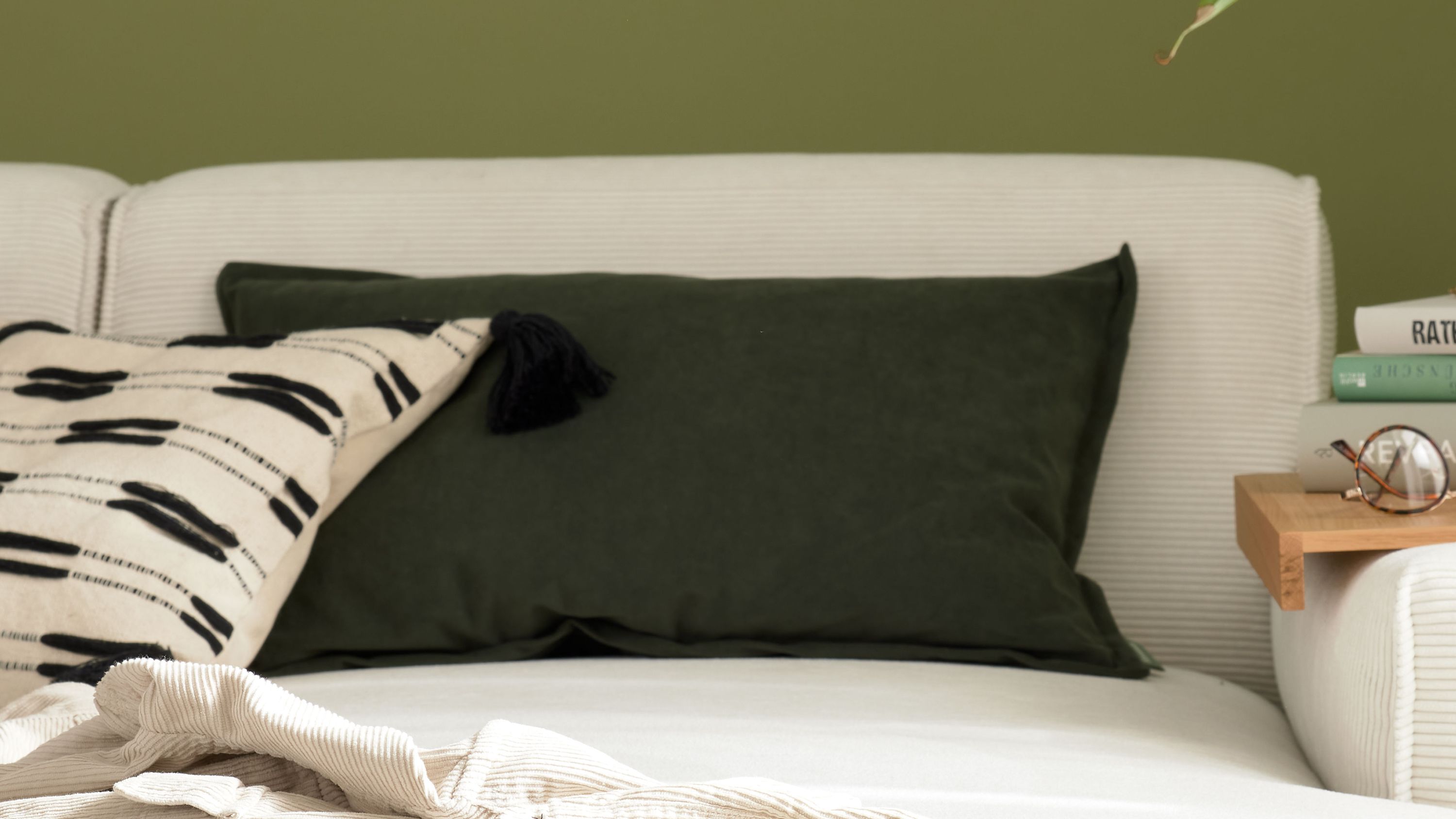 Rechteckiges Kissen mit khakifarbener Hülle auf weißem Sofa vor grüner Wand