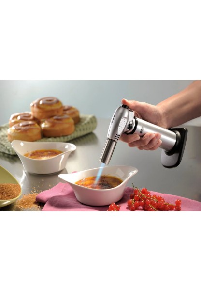 Crème Brûlée mit Küchenbrenner karamellisieren Aus schräger Perspektive: Hand karamellisiert Crème Brûlée mit Brenner, Gebäck und Johannisbeeren im Hintergrund