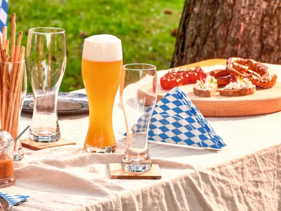 Gläserner Serviettenständer im Fokus auf gedecktem Tisch mit Getränken und Snacks im Freien, grüne Wiese im Hintergrund.