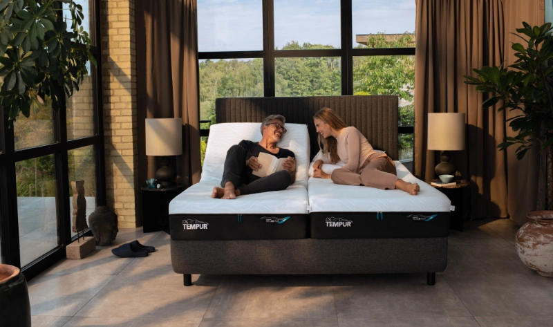 Hochwertiges TEMPUR Boxspringbett in modernem Schlafzimmer mit entspanntem Paar, natürlichen Materialien, großen Fensterflächen mit Blick ins Grüne, harmonischer Beleuchtung und komfortabler Schlafatmosphäre.