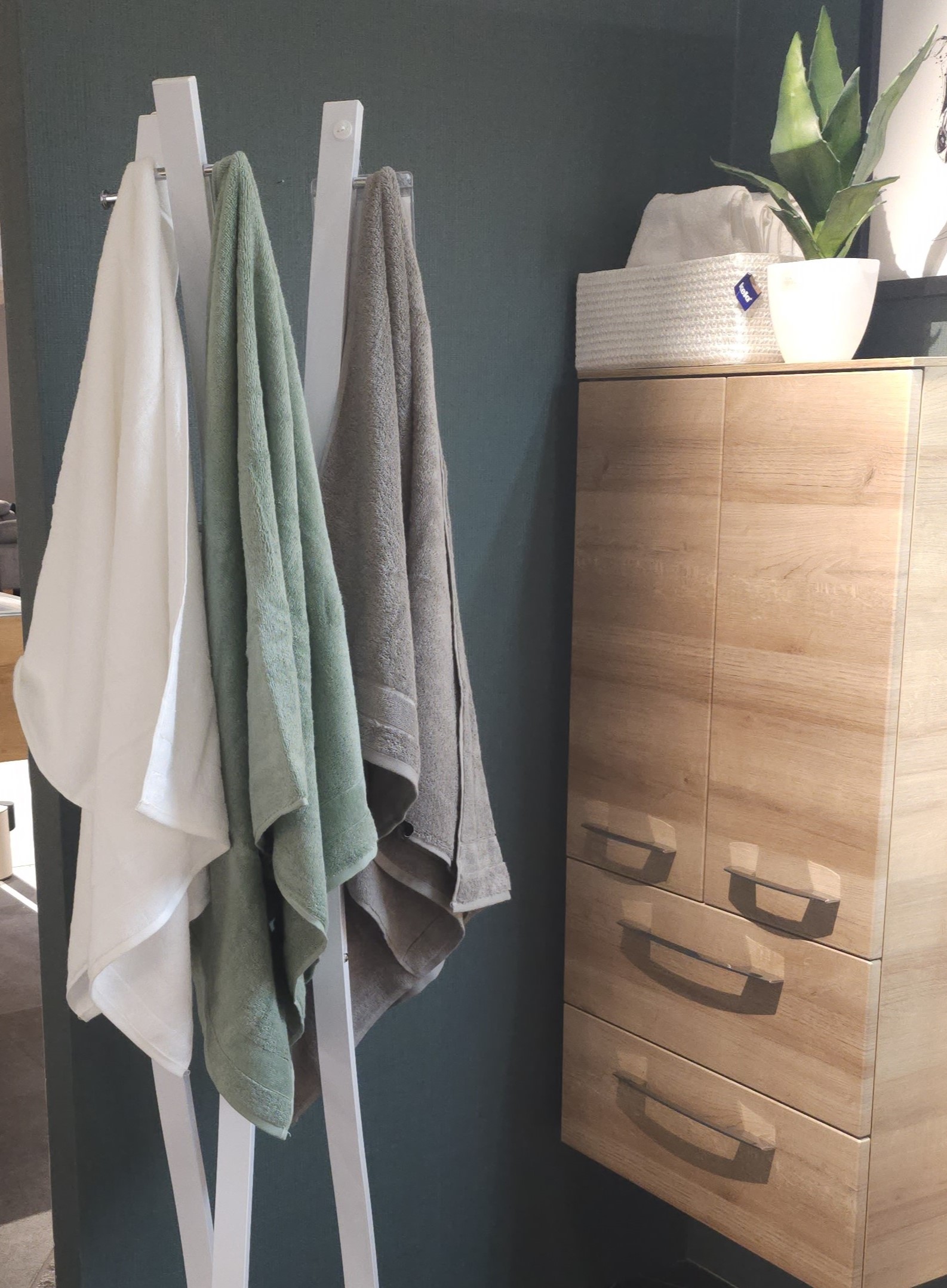 Raumecke mit 3 Handtüchern (Weiß Soft Green Grau) am Halter rechts Holzschrank mit Pflanze & weißem Behälter.