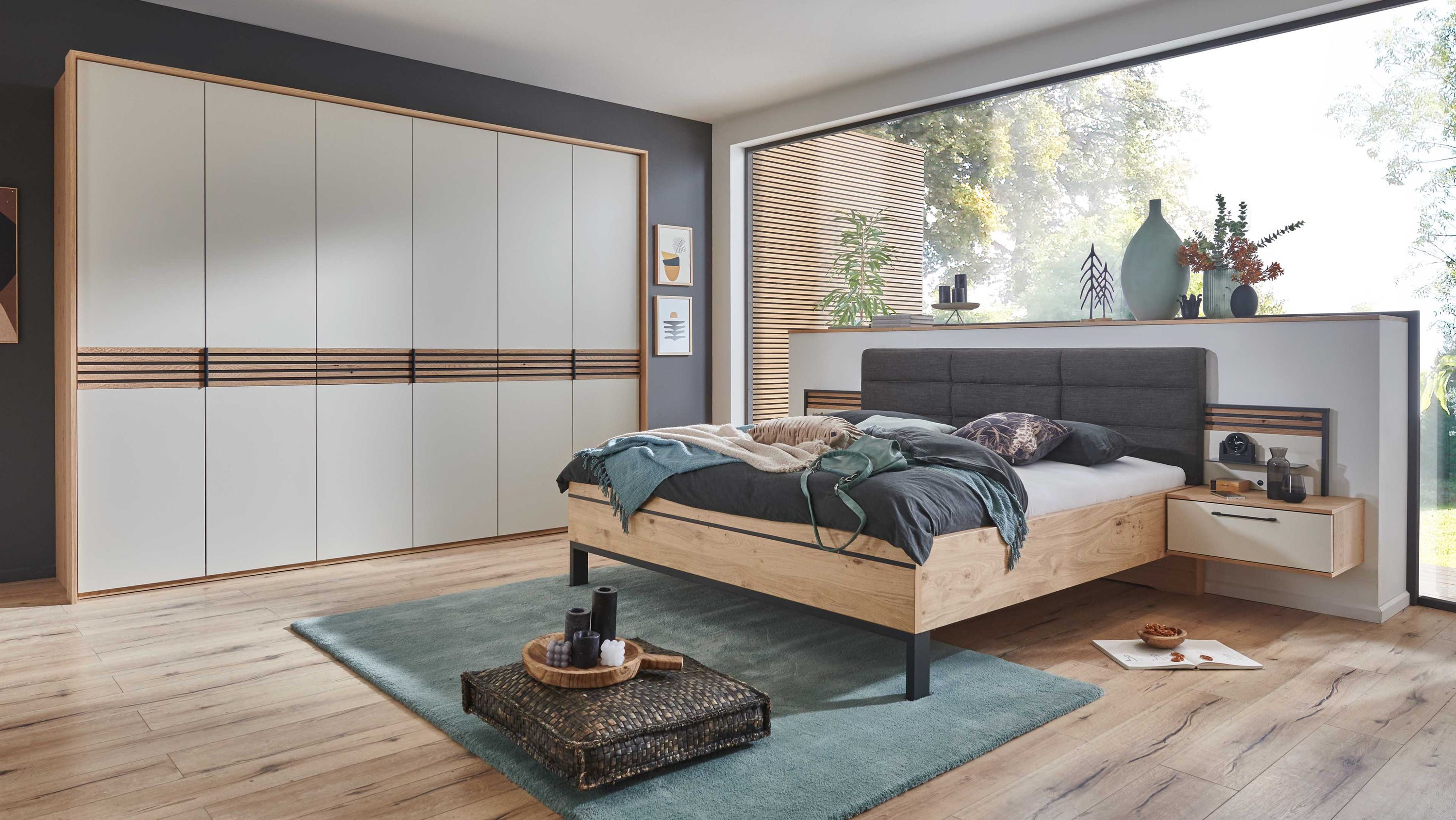 Schlafzimmer mit großem Bett aus hellem Holz schwarzem Kopfteil weißem Schrank mit Holzrahmen und blauem Teppich