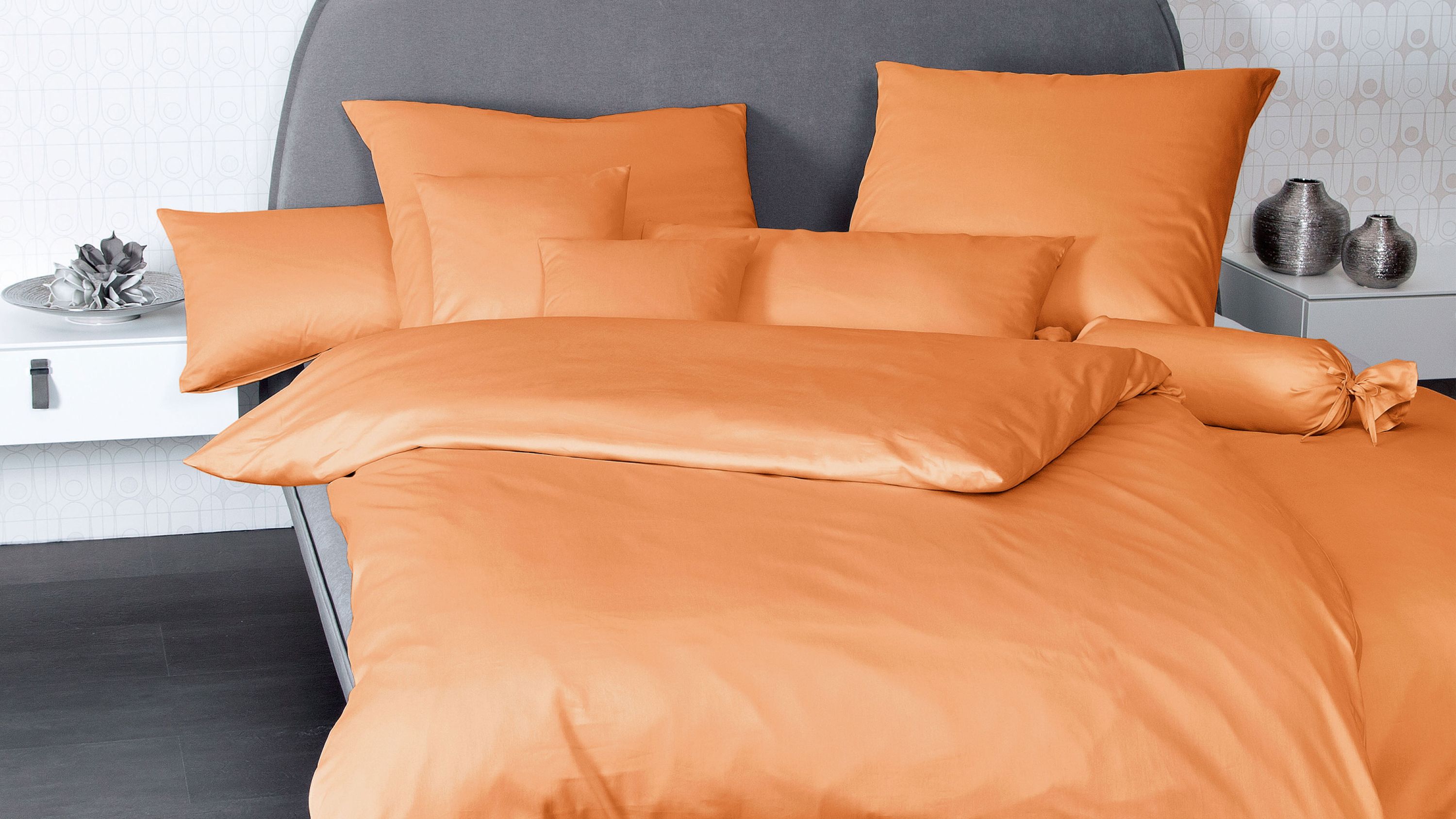 Orangefarbene Kissenbezüge in verschiedenen Formen auf einem Bett mit grauem Kopfteil