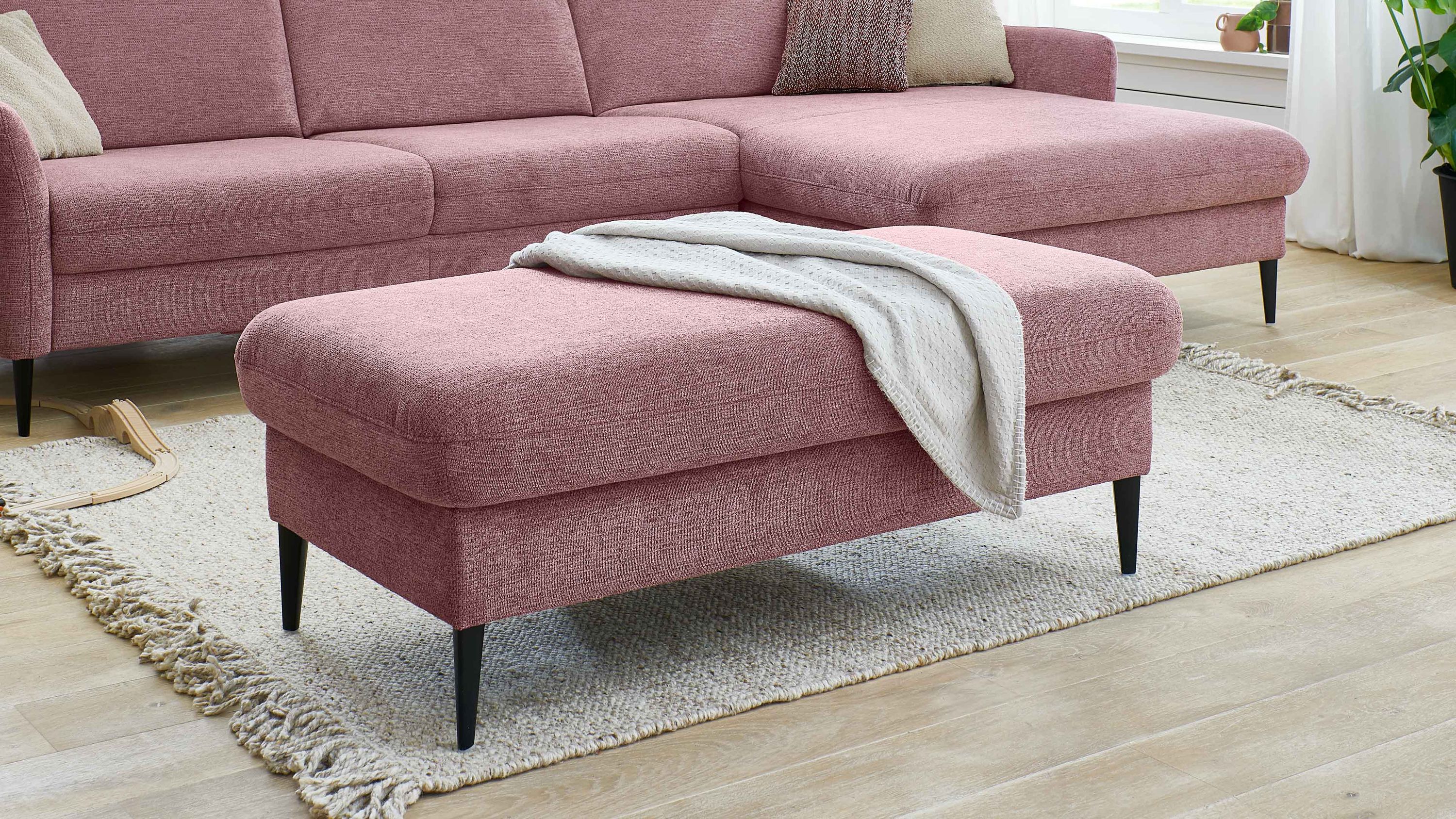 Großer rechteckiger Hocker in rosa Webstoff mit schwarzen Füßen auf hellem Teppich vor rosa Sofa