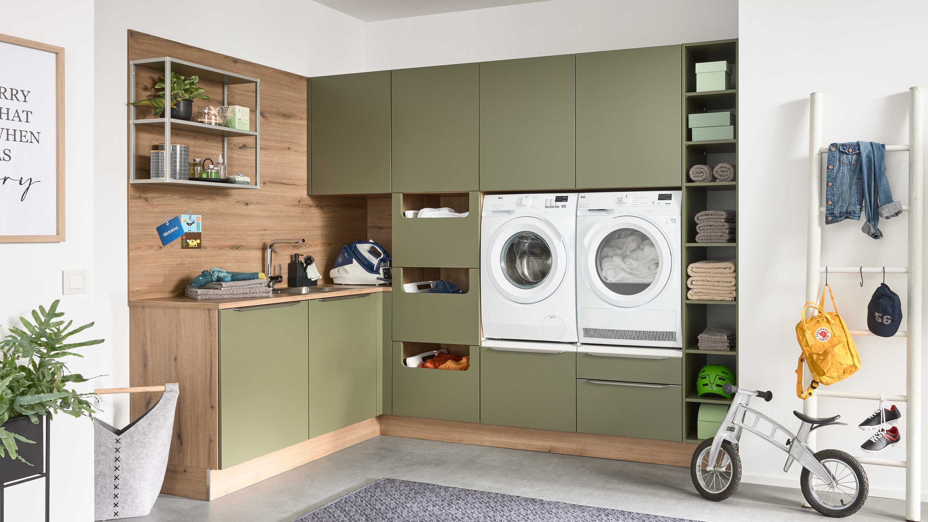 Grüne Schränke mit Holzdetails Waschmaschine und Trockner Regale mit Körben Fahrrad und Garderobe grauer Teppich