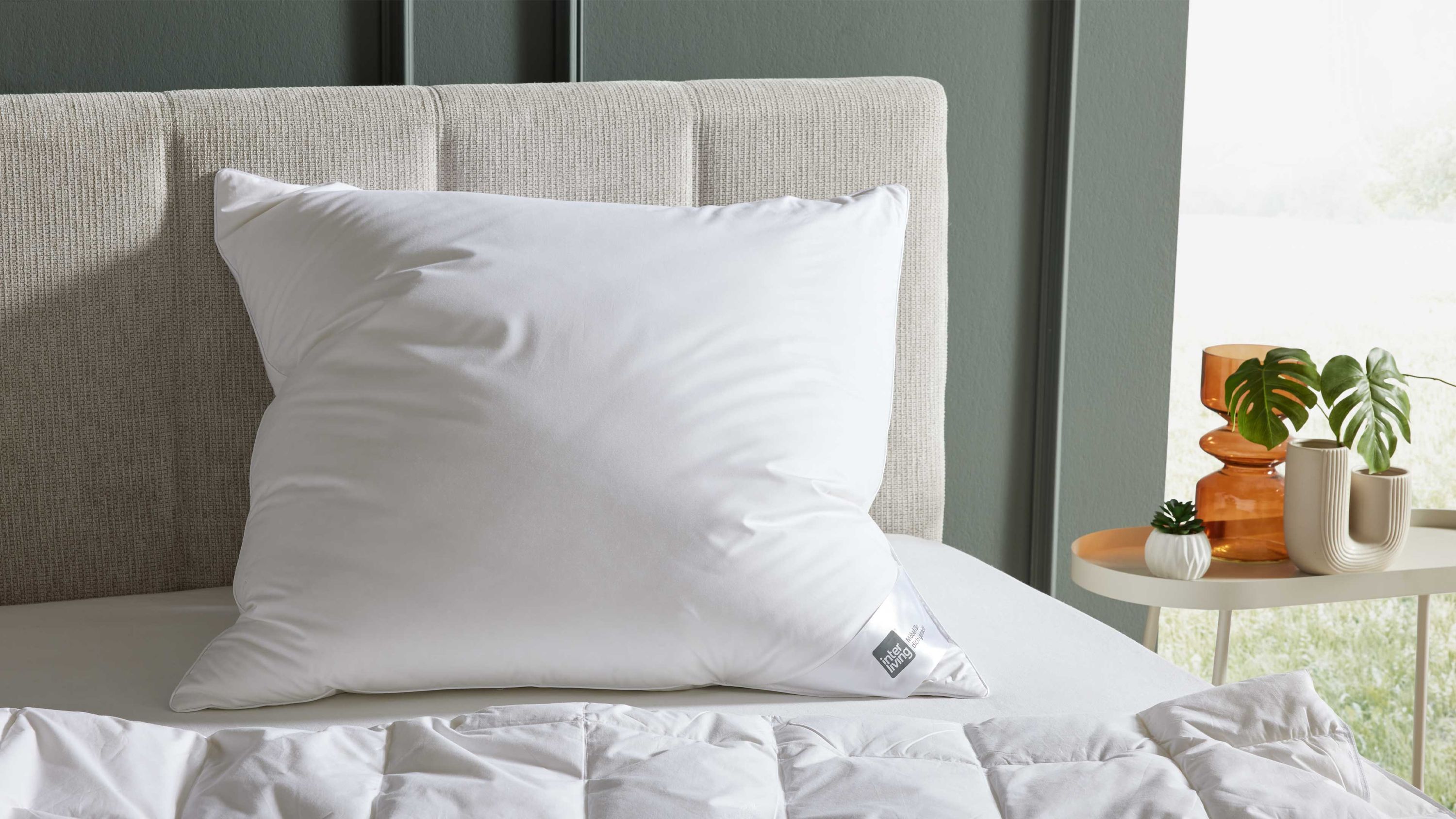 Weißes quadratisches Kopfkissen auf einem hellen Stoffbett mit beige gepolstertem Kopfteil