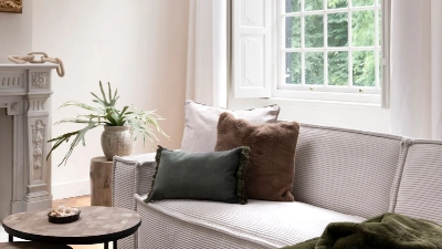 Gemütliche Sofaecke mit Ausblick ins Grüne. Helles Sofa mit Kissen und Decke vor offenem Fenster, runder Beistelltisch und Pflanze neben klassischem Kamin.