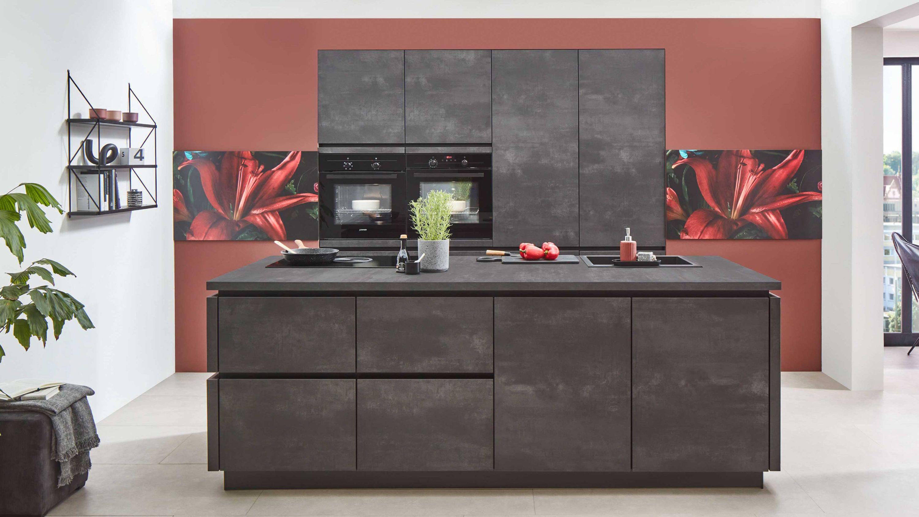 Moderne Betonoptik-Inselküche mit roter Akzentwand. Dunkle Inselküche in Betonoptik mit grifflosen Fronten Einbaugeräten und roter Akzentwand mit floralem Muster.