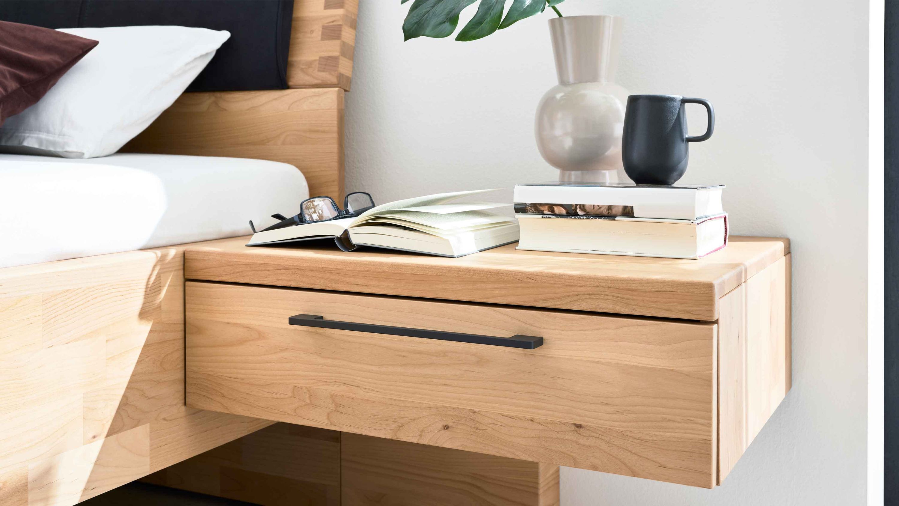 Hängende Nachtkonsole aus hellem Holz mit Schublade Hängende Nachtkonsole aus hellem Holz mit einer Schublade und schwarzem Griff neben einem Bett