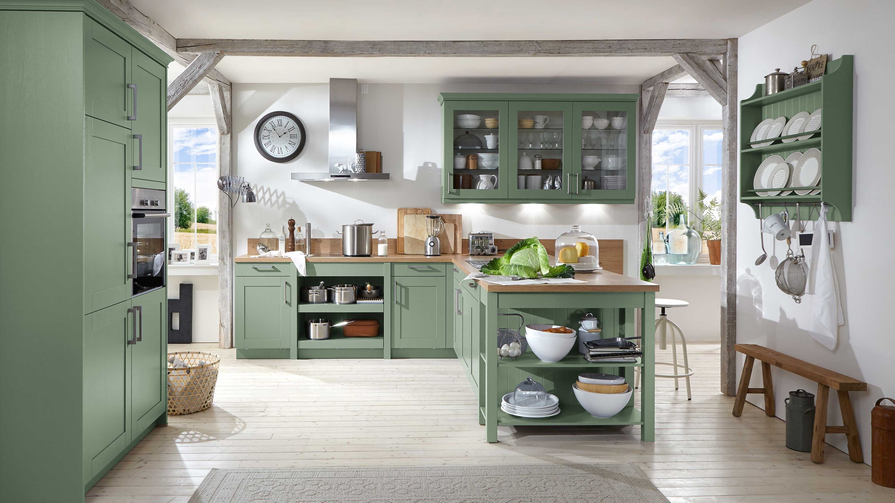 Grüne Landhausküche mit Kochinsel und Glashängeschränken. Landhausküche in Grün mit Glashängeschränken Kochinsel mit offenen Regalen Dunstabzugshaube und Holzbalken.