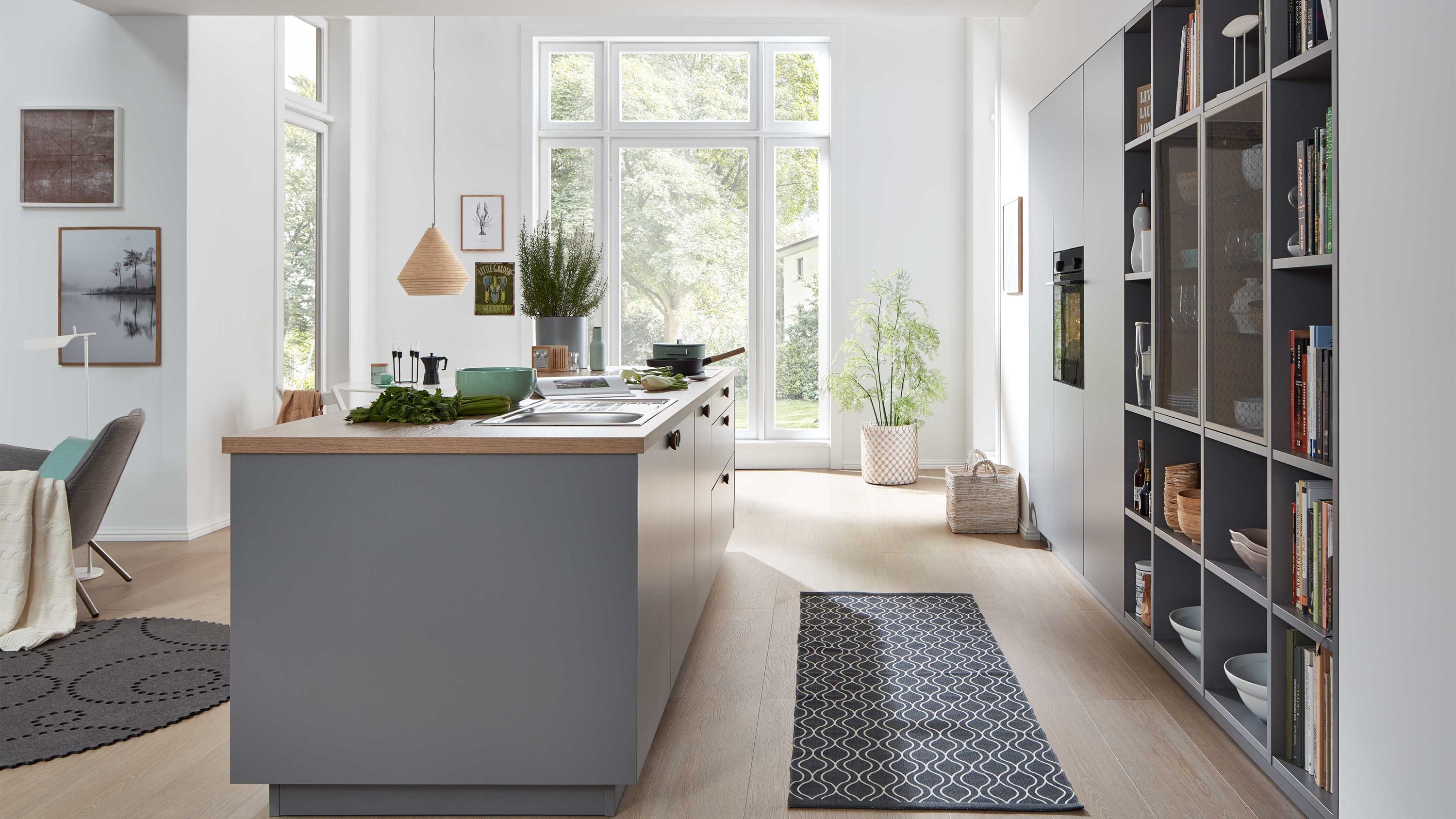 Graue Inselküche mit Holzarbeitsplatte und Regalen. Moderne graue Inselküche mit Holzarbeitsplatte hohen Regalen großen Fenstern und hellem Raumdesign.
